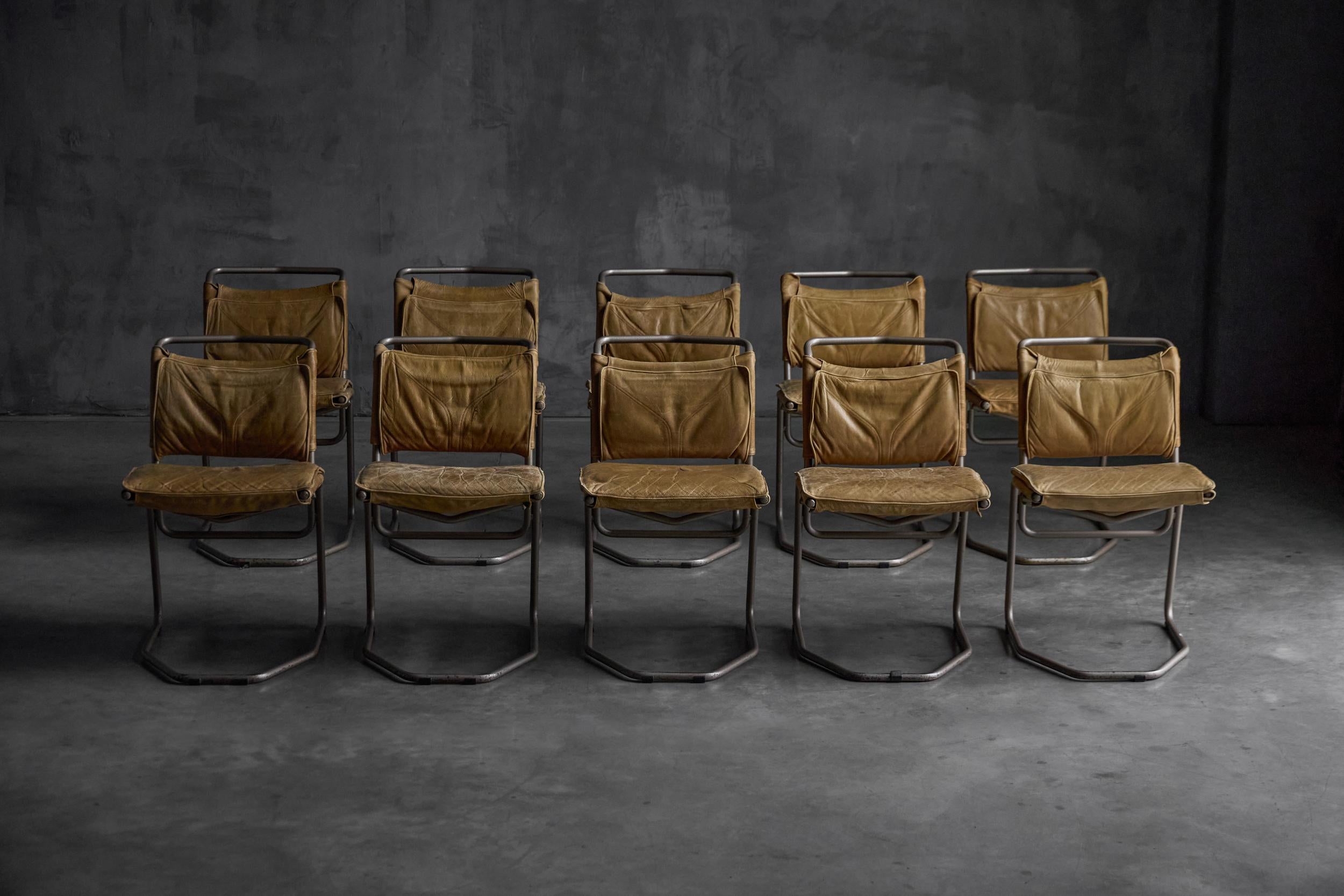 Rustic and refined dining chairs featuring original tan leather upholstery paired with minimalist steel frames. The chairs show a beautiful patina that enhances their authentic late-20th-century character. Sculptural cantilevered bases provide both