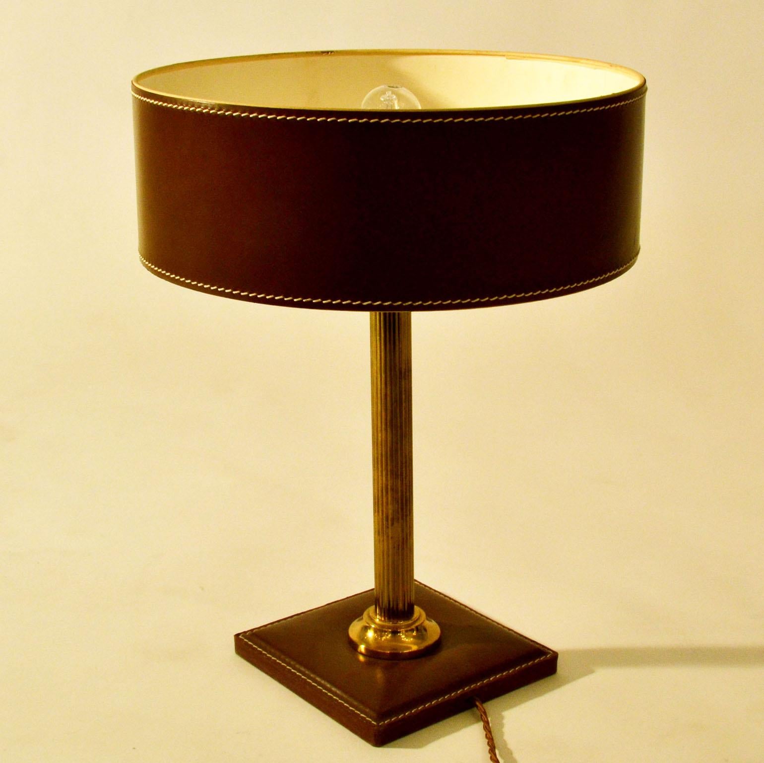 Mid brown leather table lamp with a cylinder shade and square base with detailed contrasting stitching standing on a brass stem.
Variations on this model available.