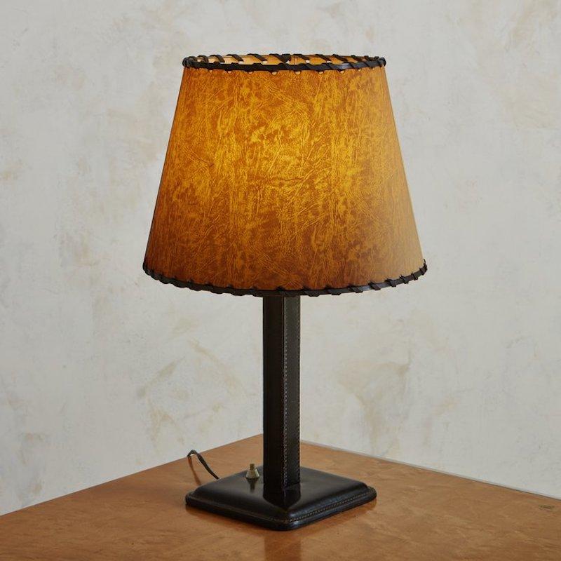 A 1940s French table lamp in the style of Paul Dupre-Lafon. This lamp is clad in stitched black leather and retains its original paper shade with stitch detailing. It has a white acrylic switch and when lit, emits a warm glow. Sourced in France,