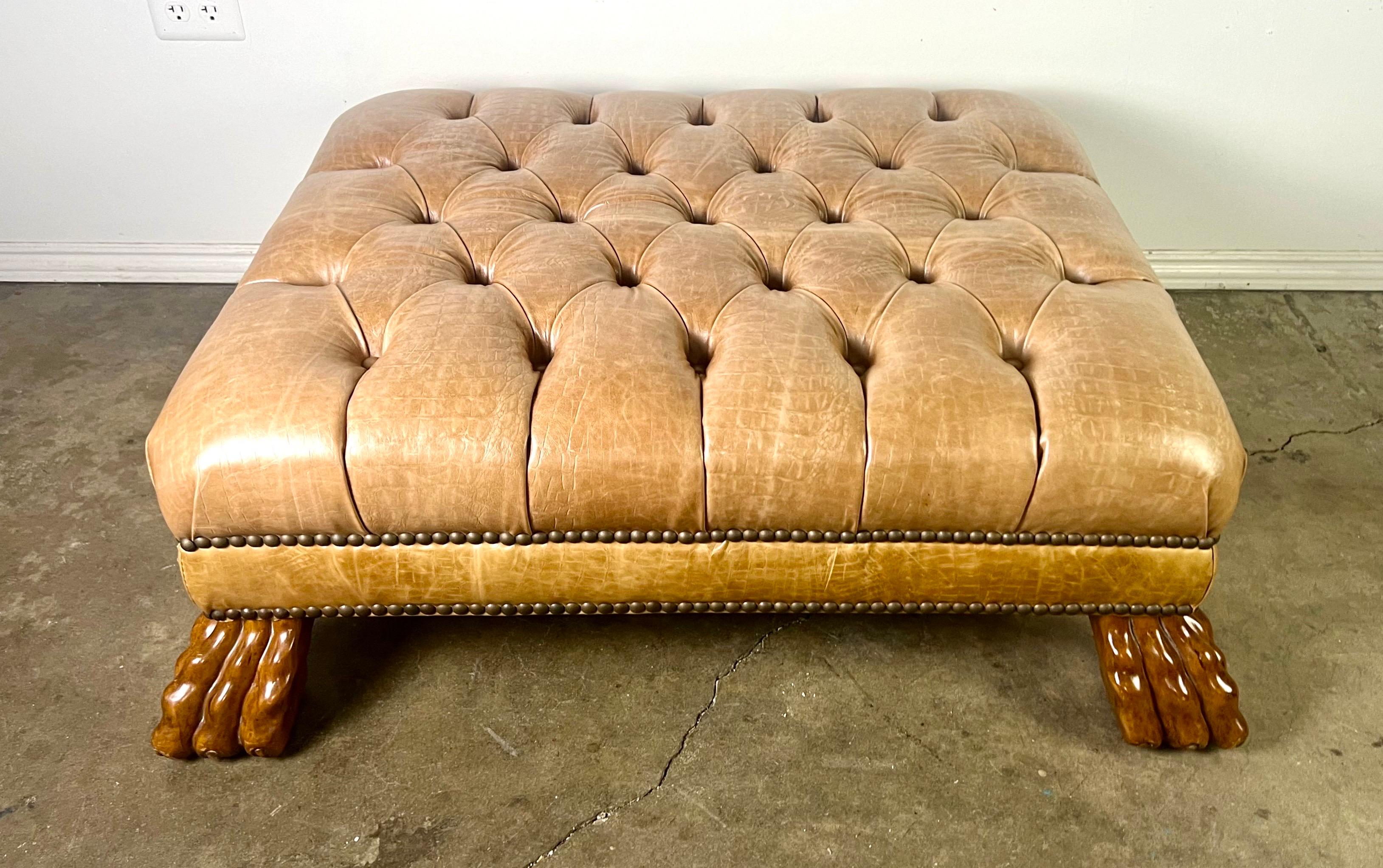 This classic English tufted embossed leather ottoman is supported by ornately carved feet, resembling lion paws-a feature often seen in Regency-style furniture.  The craftsmanship is detailed, including nailheads, emphasizing the traditional