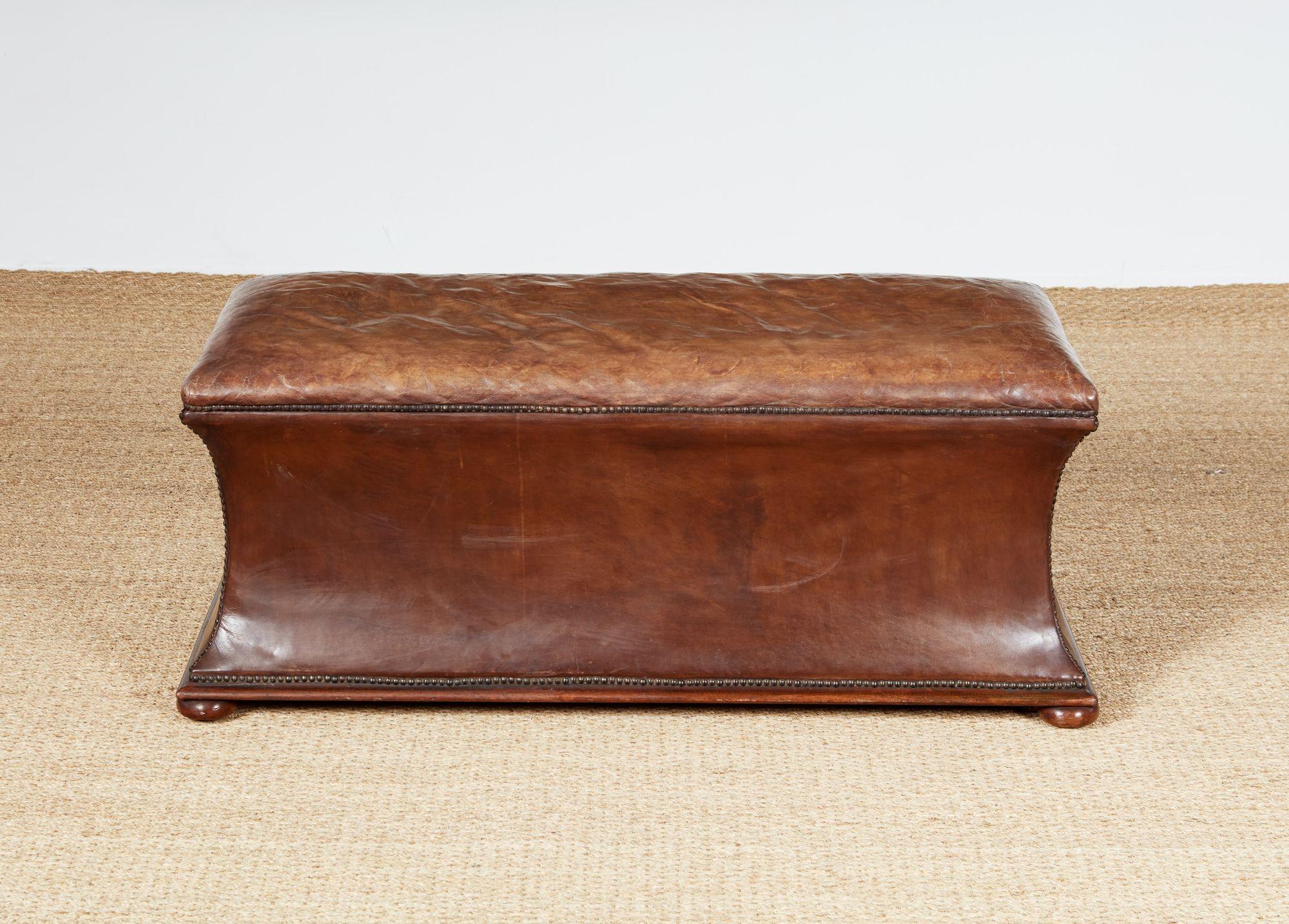Un ottoman de maison de campagne avec un dessus en cuir souple rembourré sur des côtés en cuir concave courbés vers l'intérieur et soulignés de clous en bronze au-dessus d'une base en acajou moulée sur des pieds en forme de chignon aplatis. Belle