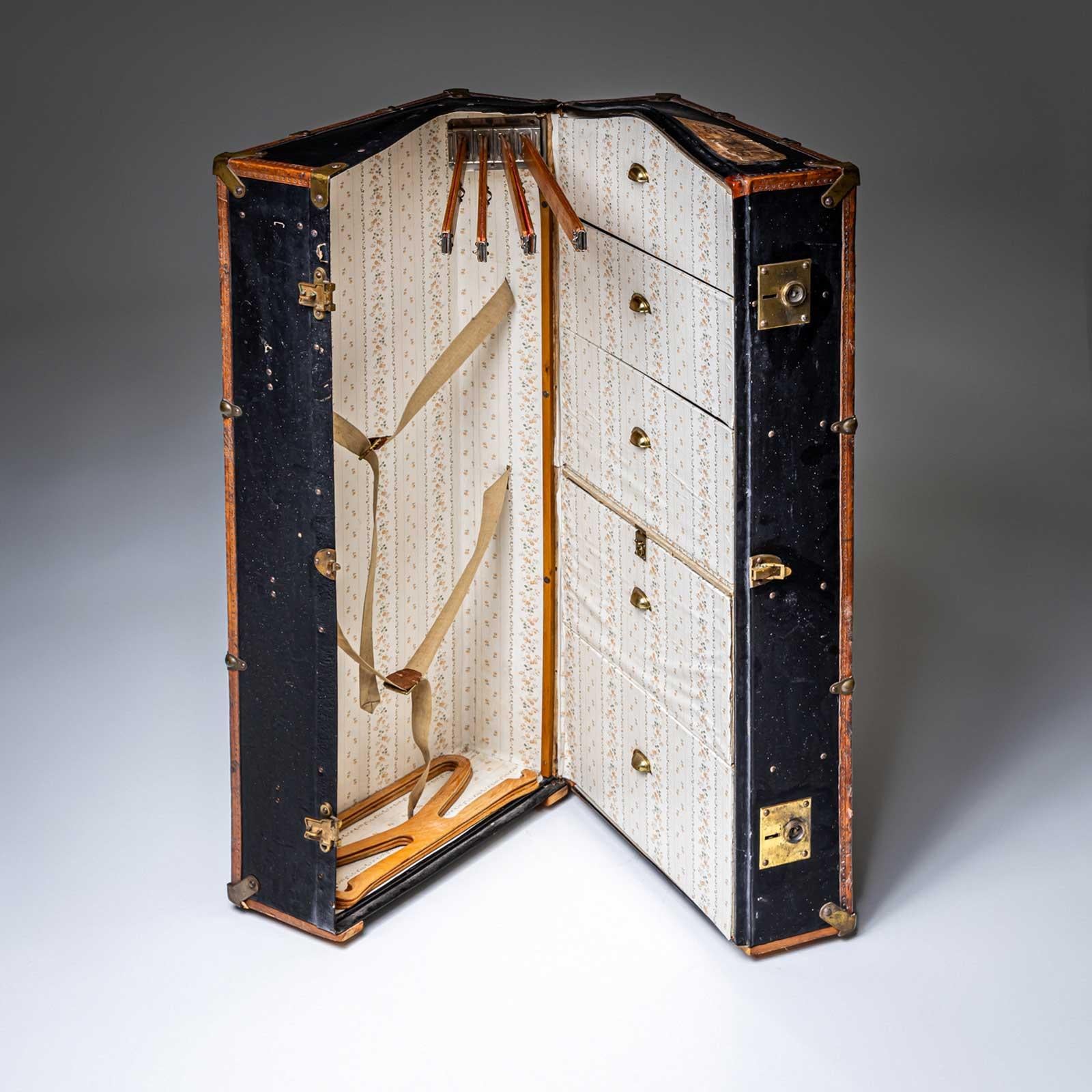 Grande baule guardaroba con rivestimento in pelle nero-marrone e accessori in ottone. L'interno del baule è rivestito con una leggera carta da parati floreale e offre un ampio spazio di archiviazione con tre cassetti, scomparti pieghevoli e