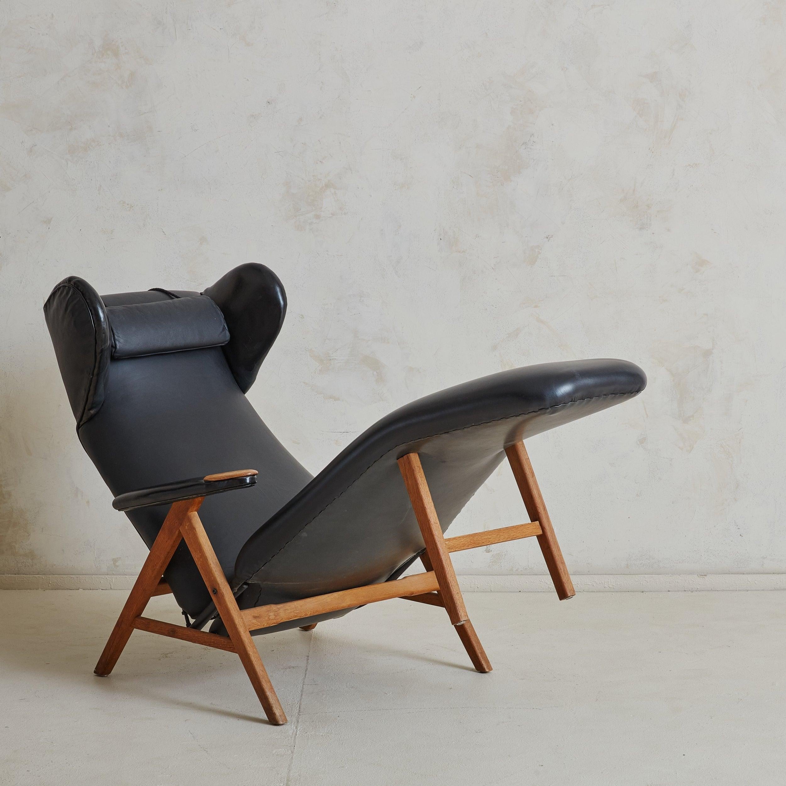 Danish Modern lounge chair features a “V” shaped honey oak frame, original black leather upholstery, padded vinyl armrests, and a removable neck pillow for added comfort. Exaggerated ‘ears’ resemble the aesthetic of a traditional wingback chair. The