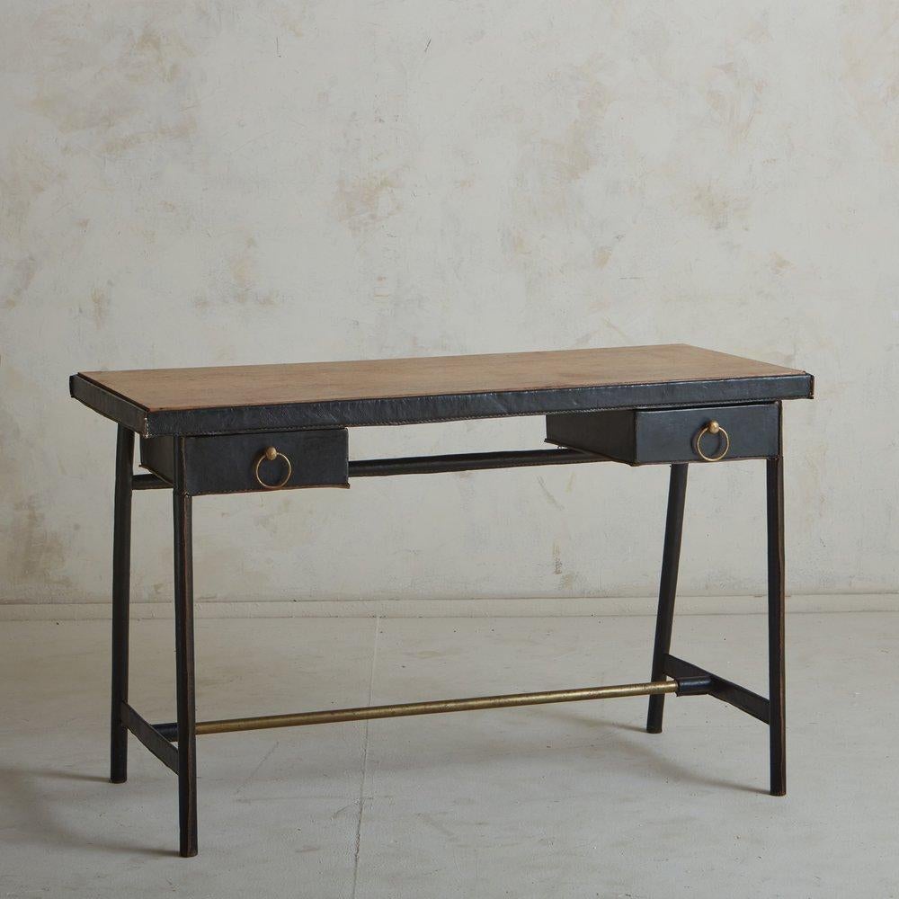 A 1950s French writing desk attributed to Jacques Adnet. This desk features a black leather clad frame with stitch detailing and an inset wood tabletop. It has two drawers with circular brass hardware and a tubular brass support bar. Unmarked.