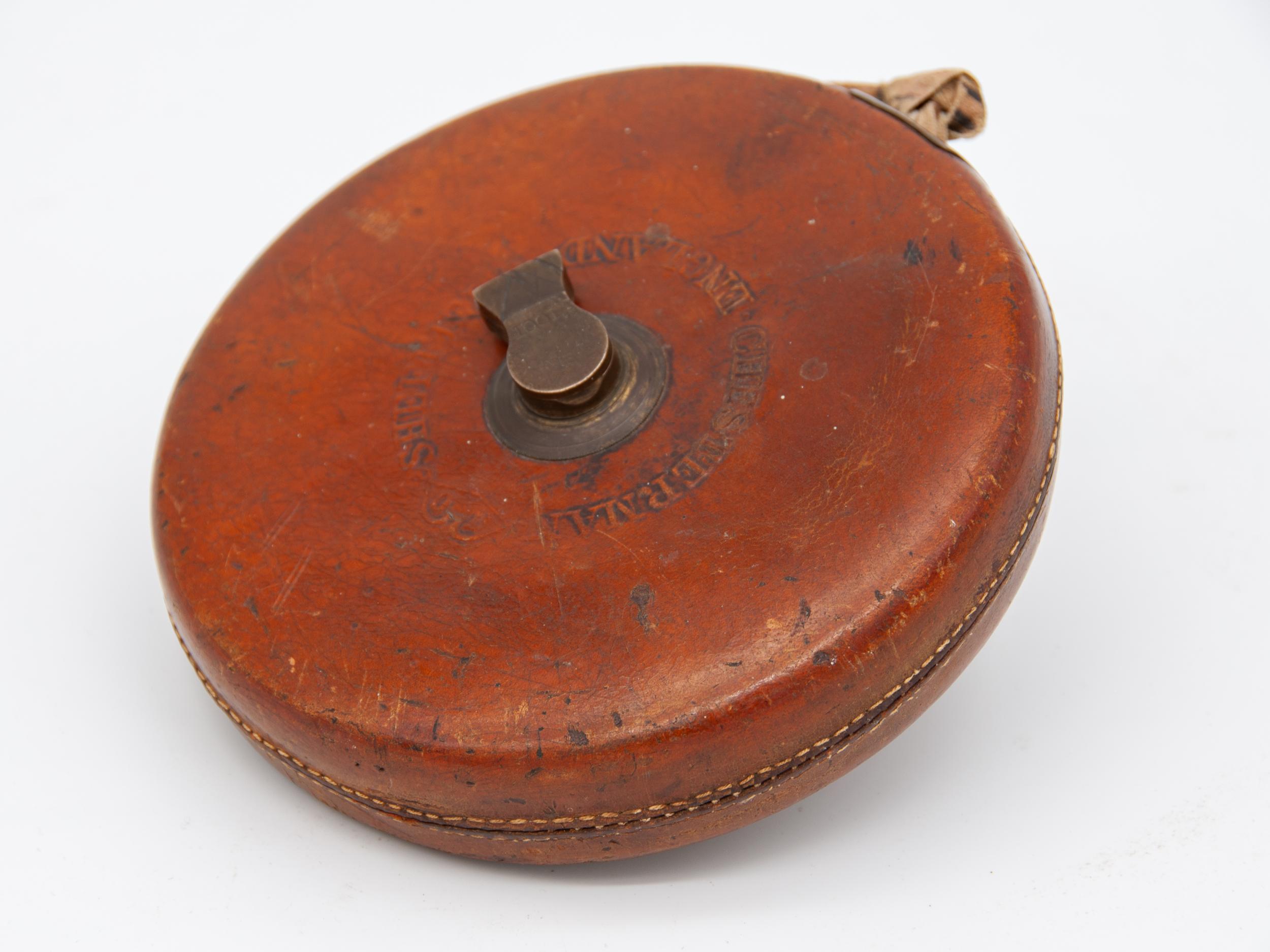Measuring tape in camel leather wrapped casing. Has a brass knob on one side to wind the tape back up. Measuring tape itself is a waxed cotton with distinct inch measurements despite being produced in the UK, circa 1900.