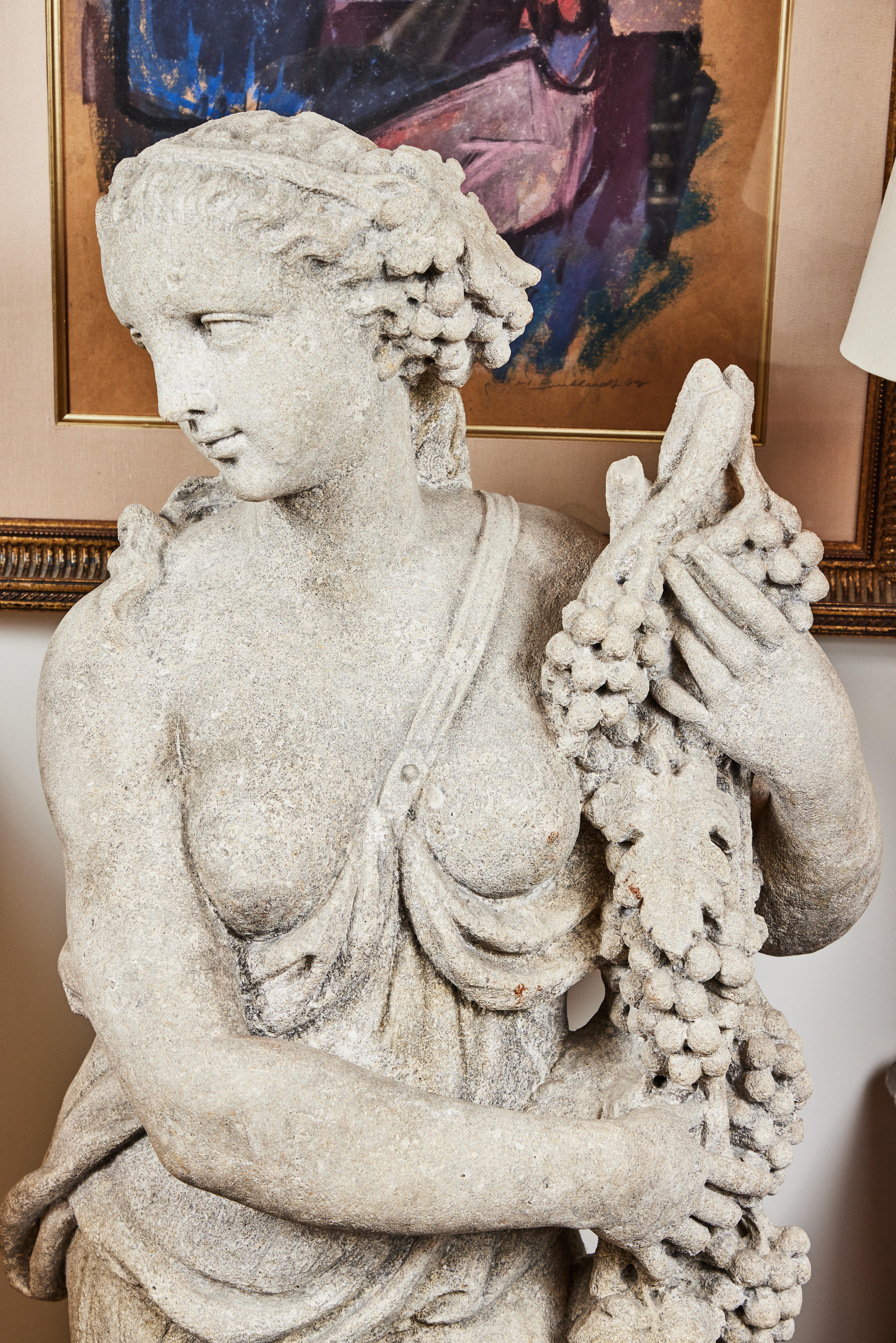 A pair of circa 1920, graceful, Italian, limestone, left-and-right nymph sculptures in cascading drapery, possibly allegories of Spring and Summer. Each cradling a garland of flowers and a grape vine, respectively.