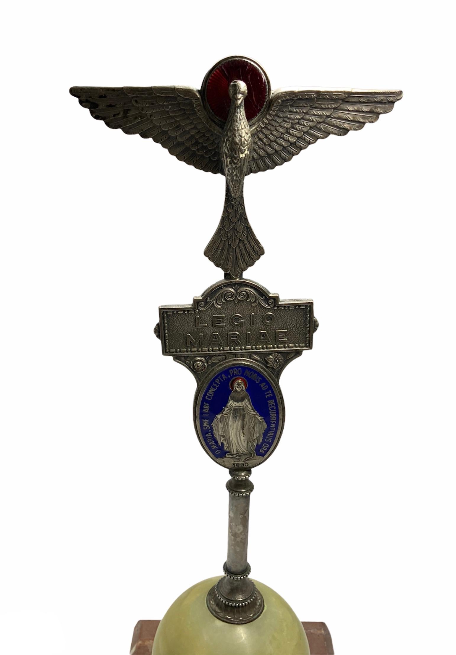 This is a Legio Mariae statue composed of an open winged silver tone dove (representing the Holy Spirit) with red enamel halo. Below it, there is a silver plaque with the name Legio Mariae written and then an oval silver and blue enamel cameo of the