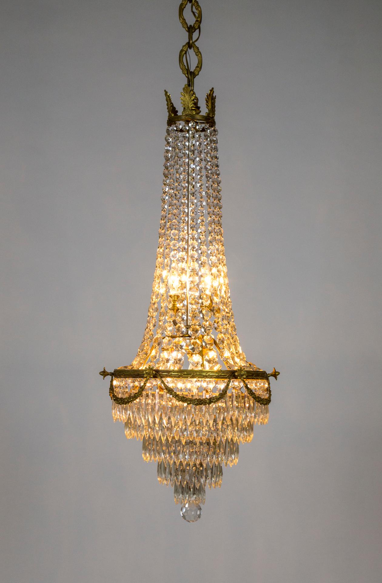 An oversized, slender, neoclassical style, gilded bronze and crystal chandelier, in tent-and-bag shape, with reeded ring and foliate swag detailing, and strands of heavy, crystal beaded drapes from ring to crown. It has 5-light, 3 illuminating the