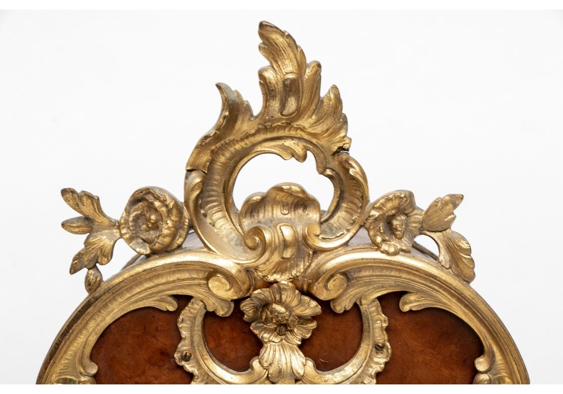 A mantel clock in reddish- brown figured wood veneer with dore mounts in the baroque style. The dore dial with enamel numerals, the case with dore scrolled acanthus leaf frame with cartouche and floral crest. A glass cartouche on the front to view