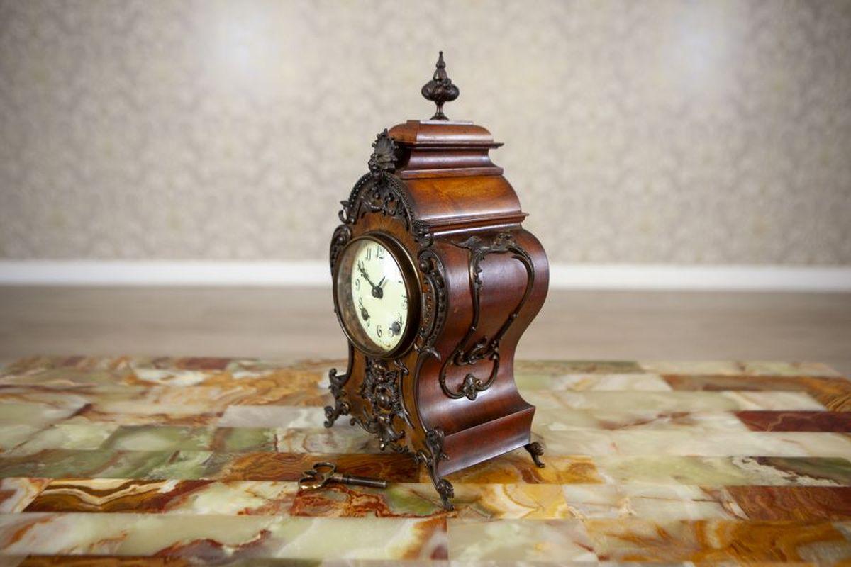 Lenzkirch Mantel Clock Circa 1895-1900 in Walnut Case

A Lenzkirch mantel clock, dated circa 1895–1900 (based on the serial number visible on the back of the clock). The clock features a wooden case adorned with floral brass decorations and rests on