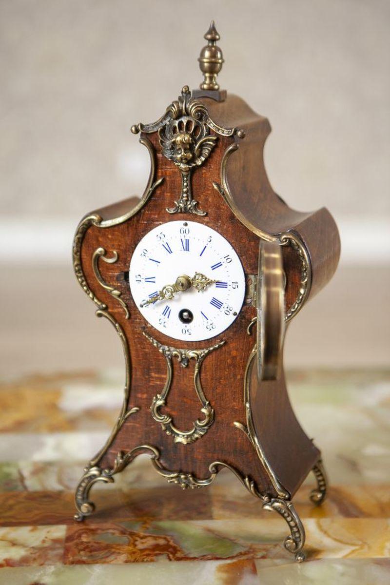 Lenzkirch Mantel Clock from the Early 20th Century in Walnut Case

Lenzkirch Mantel Clock, dated around 1903-1905. The piece features a wooden case adorned with brass fittings, decorated with floral motifs, and a cherub face, resting on four small