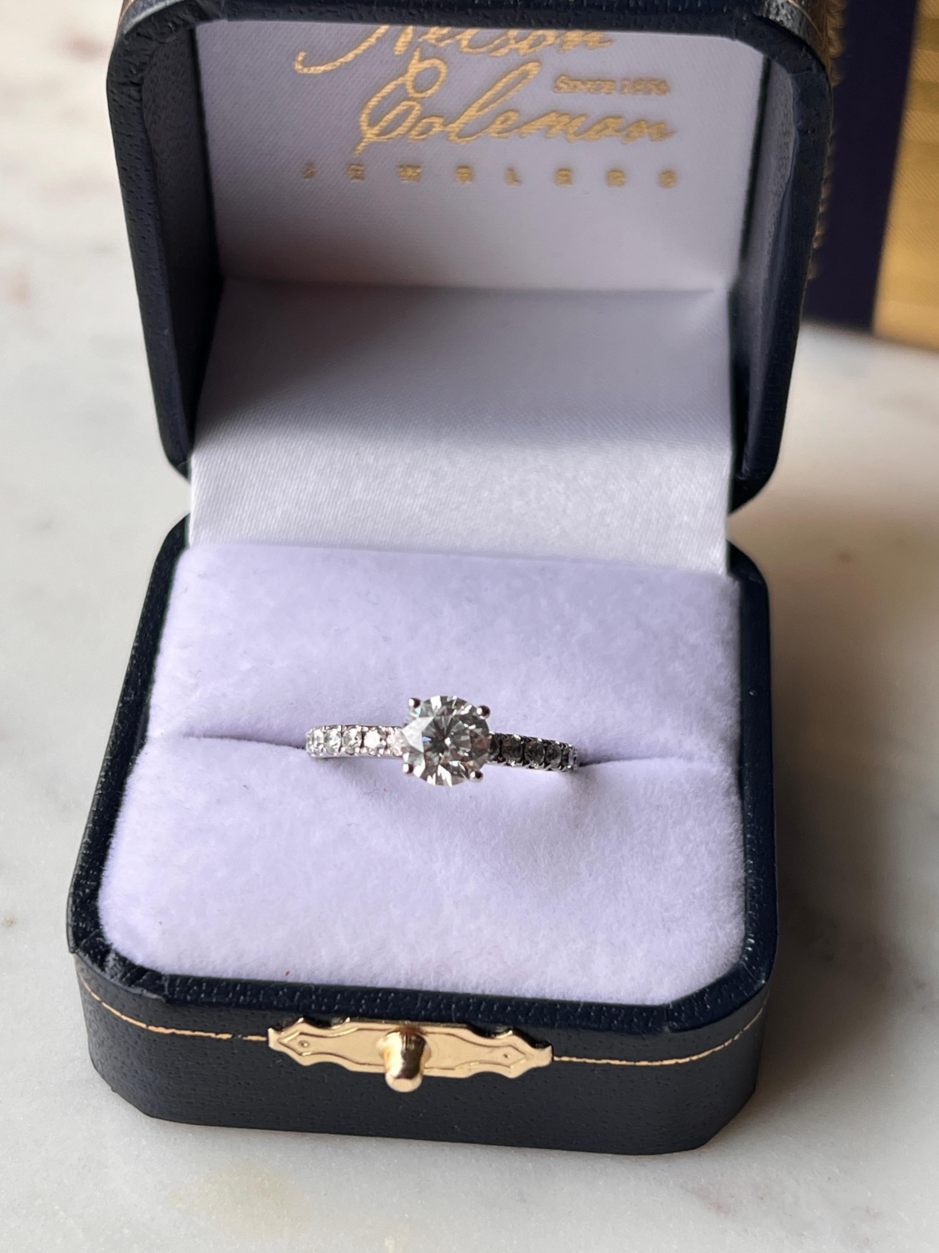 Questo squisito anello di fidanzamento in oro bianco 14K di proprietà mette in mostra una brillante pietra centrale di diamante tondo Leo da 0,98 carati di eccezionale colore F e purezza SI2, accompagnata da scintillanti accenti di diamante sulla