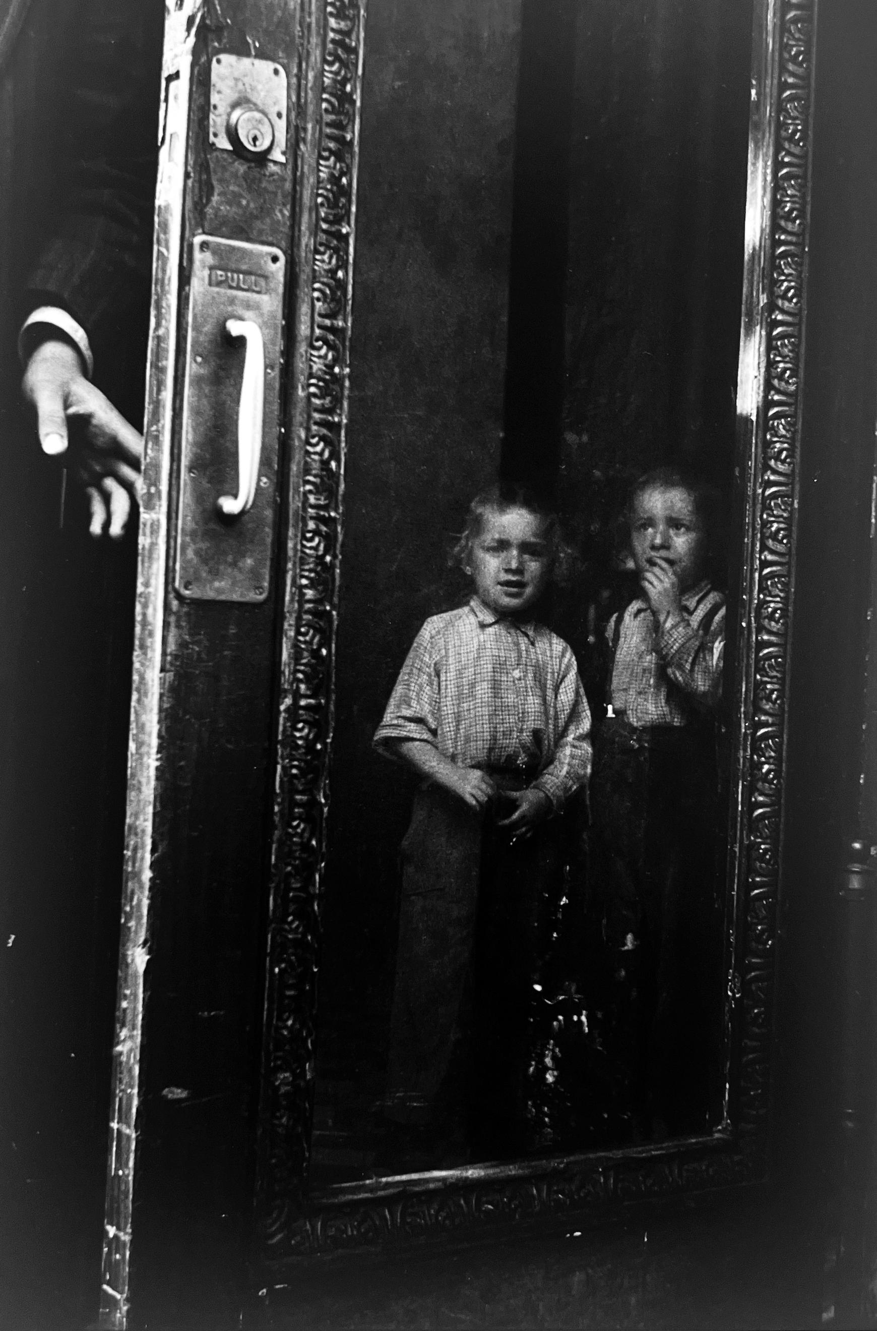 Yeshiva Boys, 1954 is a black-and-white photograph of two young studious boys behind a glass door, an entrance to a Brooklyn brownstone in New York City. A gelatin silver lifetime print, 15.5” x 12