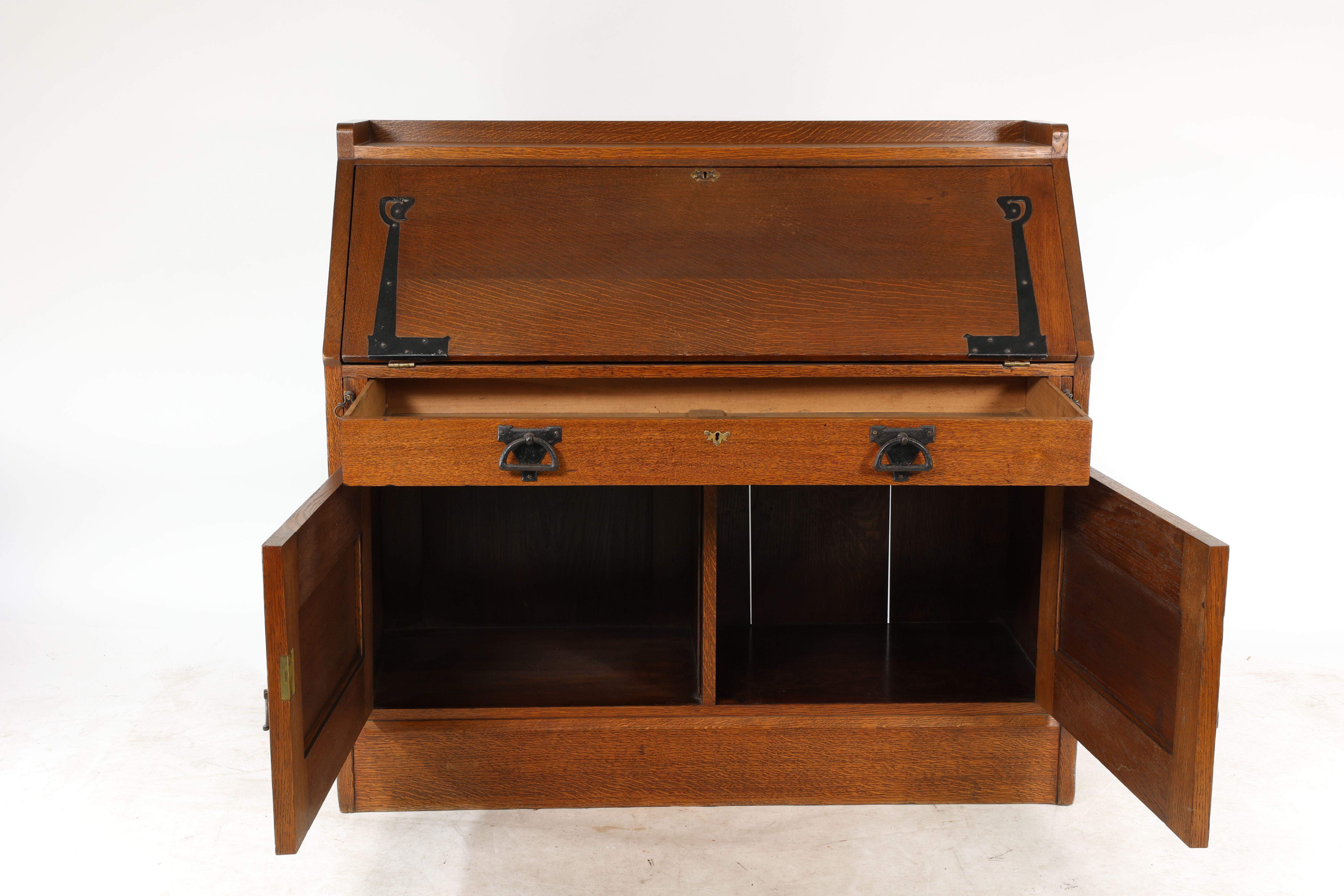Liberty and Co. Leonard Wyburd. An oak bureau with drop down writing area held in place by slides each side of the large drawer with a large double cupboard below all with stylised iron strap hinges.