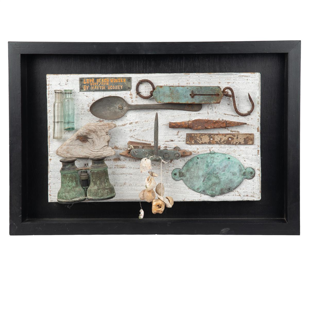 Eine gerahmte Collage aus Treibholz, Glasflaschen, Kieselsteinen und Metallgegenständen, darunter ein Löffel, eine Waage mit der Aufschrift 