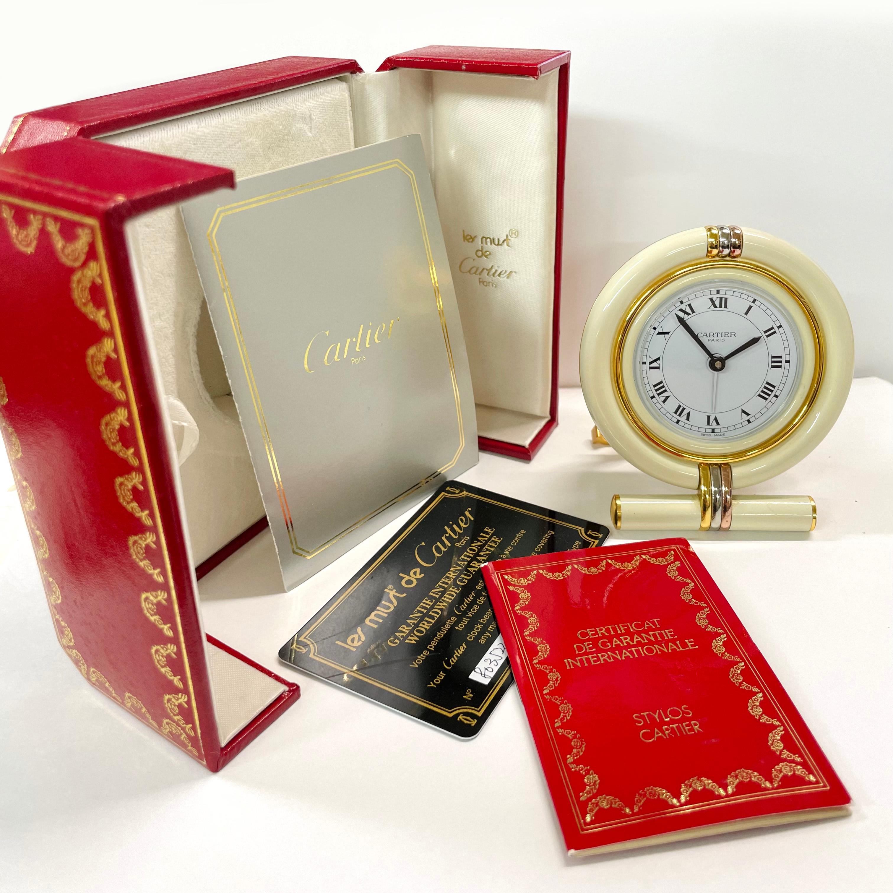 Les Must de Cartier Paris Enamel Gold Plated Travel Clock with original case and manual. This beautiful and practical travel alarm clock has a quartz Swiss movement with Cartier C's on the bottom of the stand and cream colored overall enamel with