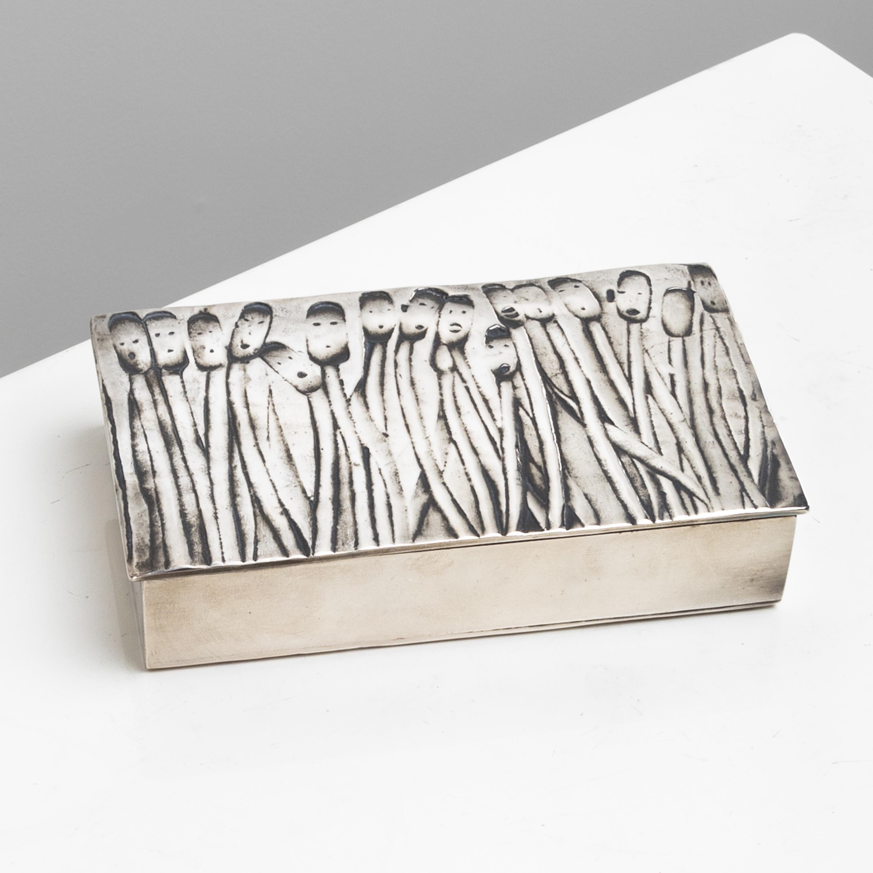 “Les Roseaux à visage humain” (“Reeds with Human Faces”) is a double-sided silvered bronze box by Line Vautrin.
The front side representing reeds (rushes) in motion, with their flowers replaced by human faces.
The engraved underside of the box