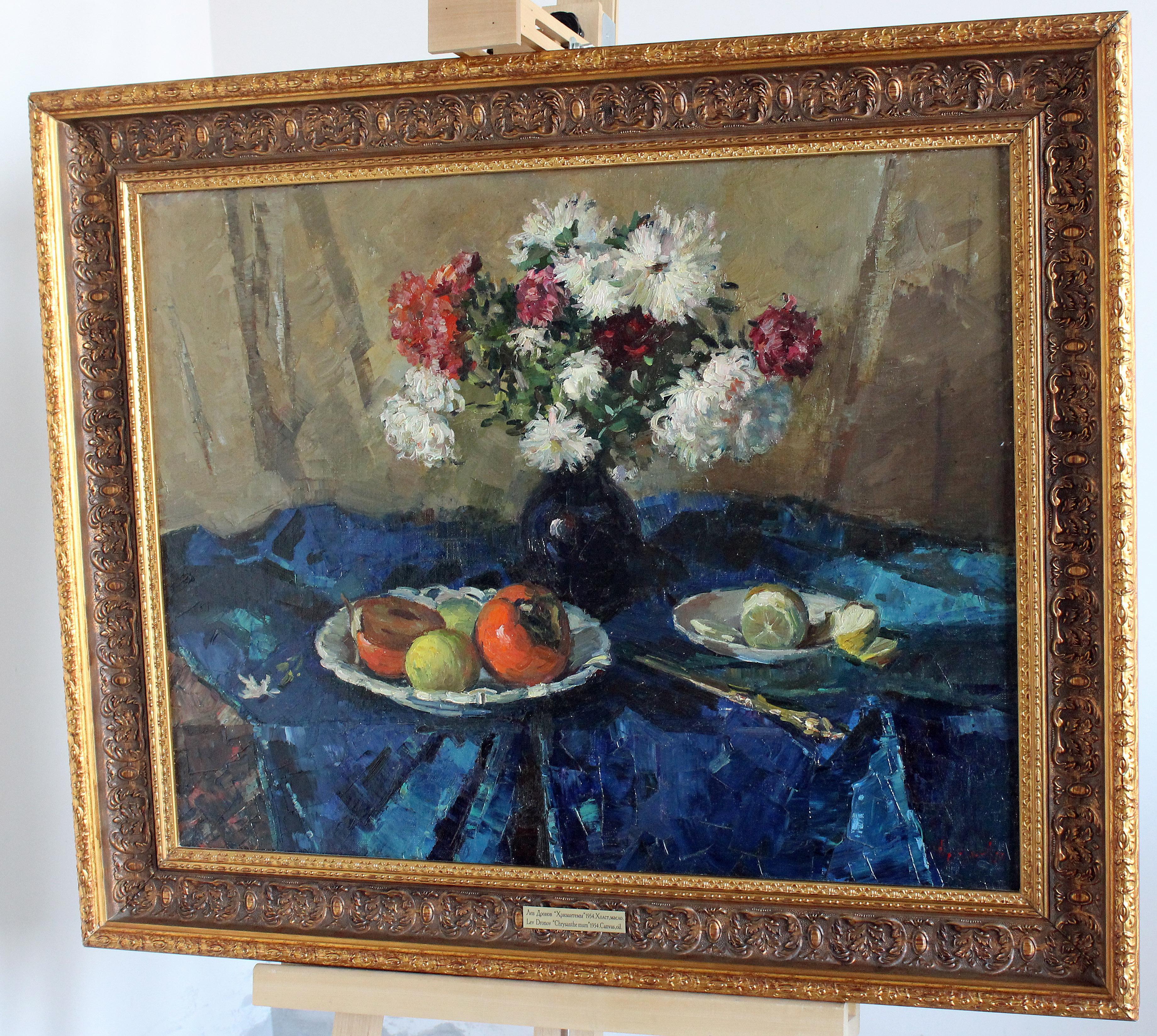 Sur le plan de la table, recouvert d'une draperie bleue, il y a un vase avec des chrysanthèmes de couleur blanche et violette, pratiquement au centre. Le fond est une toile de lin grisâtre neutre. Les coupes de fruits sont au premier plan.
Dans