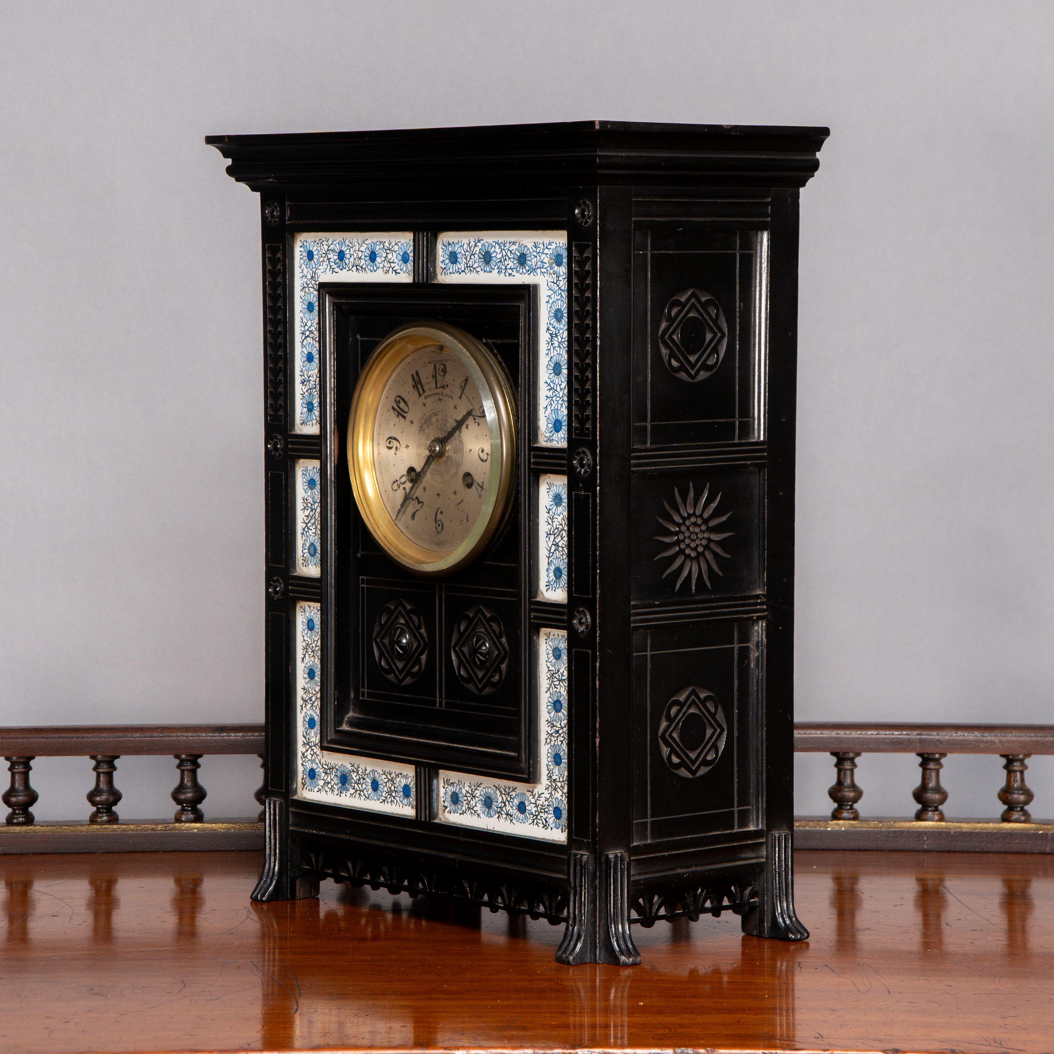 An Aesthetic Movement ebonized clock with finely carved decoration, the clock movement is by Edwards and Sons with a pair carved devices below and a blue and white floral ceramic border. The finely fretted decoration to the base is identical to the