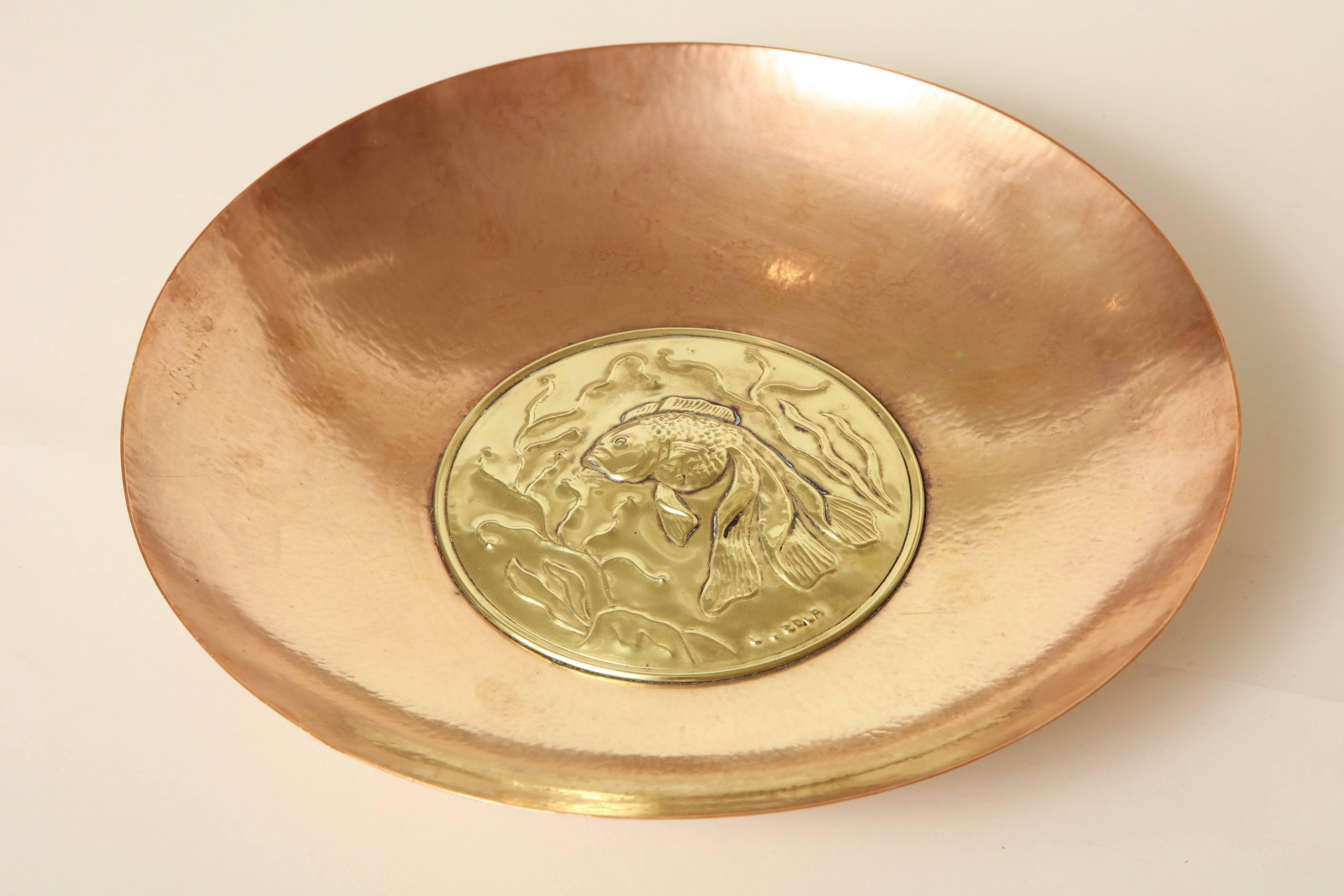 Measures: 11 ¾” diameter; 2 ¼” deep
medallion 5 3/8” diameter.

Circular copper coupe with central brass fish design.
Impressed Zola.