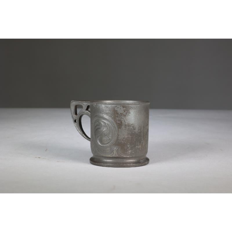 Liberty and Co. Estampillé Made in England Tudric 0358. Mug de baptême en étain de style Arts and Crafts, avec une décoration en forme de tourbillon et une anse stylisée et percée.
