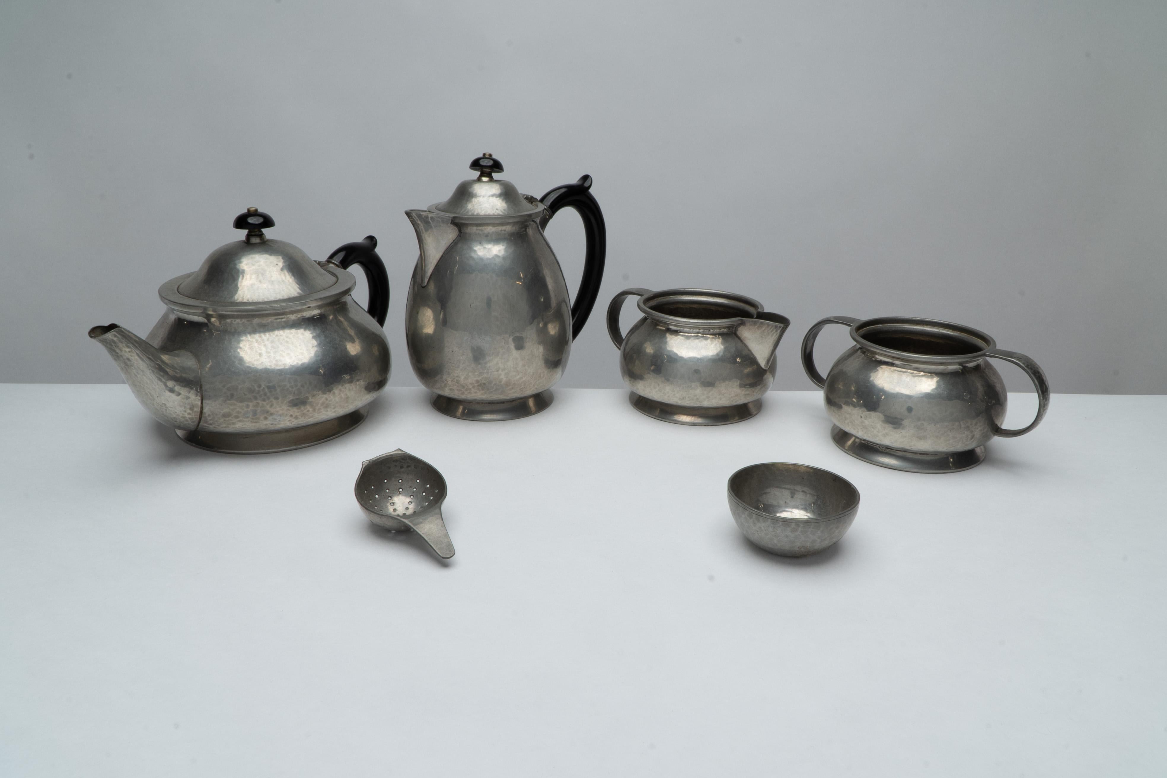 Liberty and Co. An Arts and Crafts six piece pewter tudric tea set, consisting of a teapot, water jug, milk, sugar and a tea strainer with base. Stamped to the base, '6, Tudric, English Pewter, Liberty & Co, 01075, two pints'. Dimensions: Teapot: H