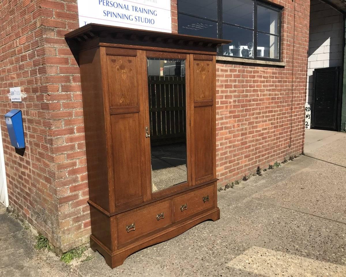 Liberty & Co, attributed. An Arts & Crafts double oak wardrobe with flaring cornice, centre by a full length bevelled mirror door with stylised floral inlays and fine line inlays and two large deep drawers to the base with hand beaten copper handles.