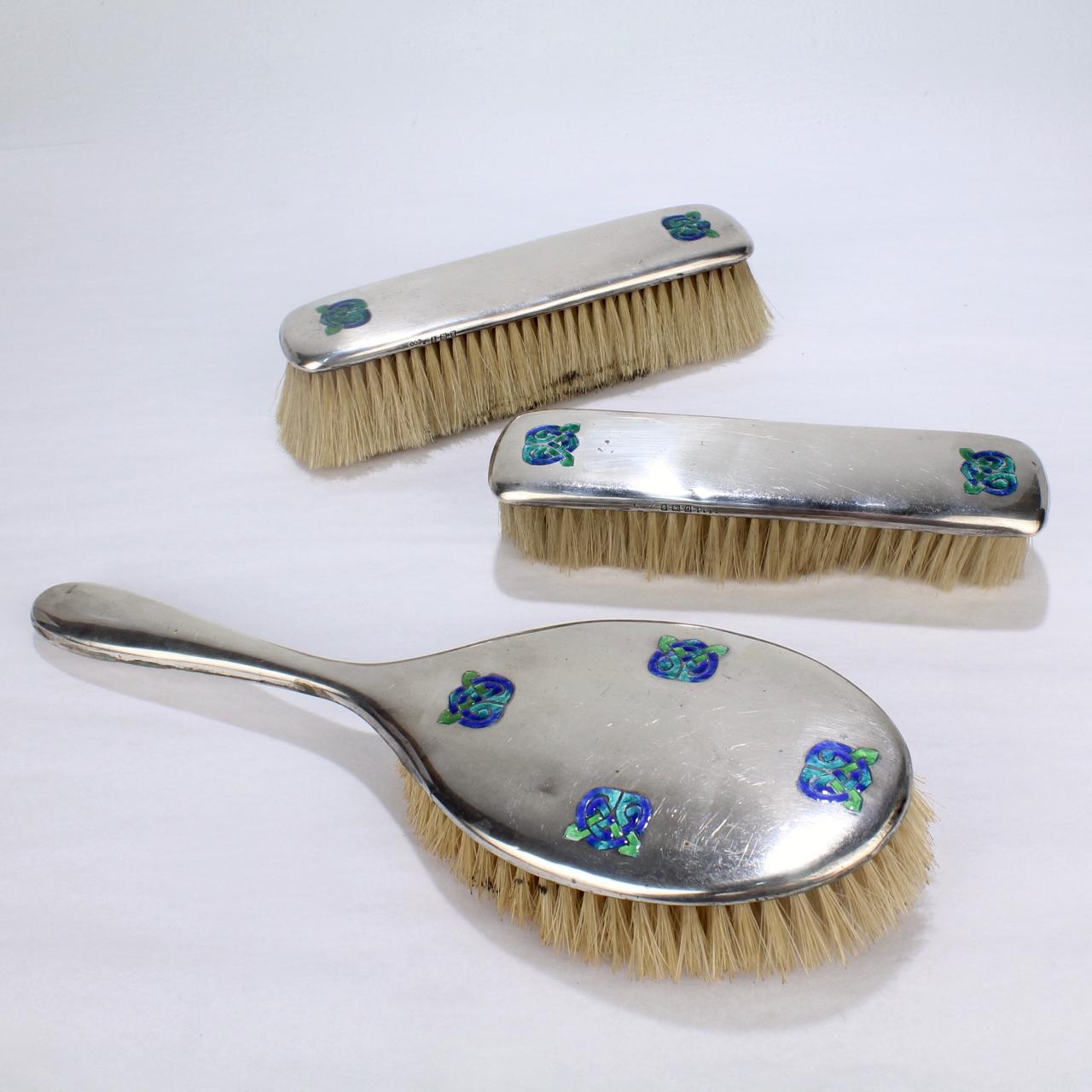 A wonderfully rare Art Nouveau Cymric pattern sterling silver partial dresser set including a hand mirror and three brushes.

The Cymric pattern was designed by Archibald Knox for Liberty and Co. and features green & blue enameled Celtic knot