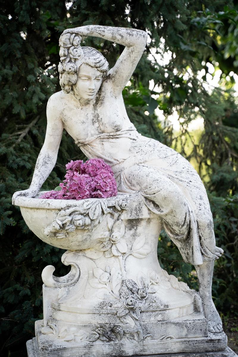 Una splendida scultura decorativa in graniglia di marmo, realizzata durante l'apice del periodo Liberty (Art Nouveau) negli anni Trenta. La figura femminile, graziosamente appoggiata su un bacino ornato di fiori, incarna perfettamente l'eleganza