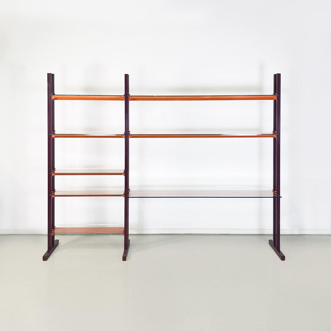 Metal and glass bookcase model Caos San with a structure composed of three burgundy-colored metal uprights, with orange metal and glass shelves of different sizes between the first and second bays, where in addition to a greater width of the same