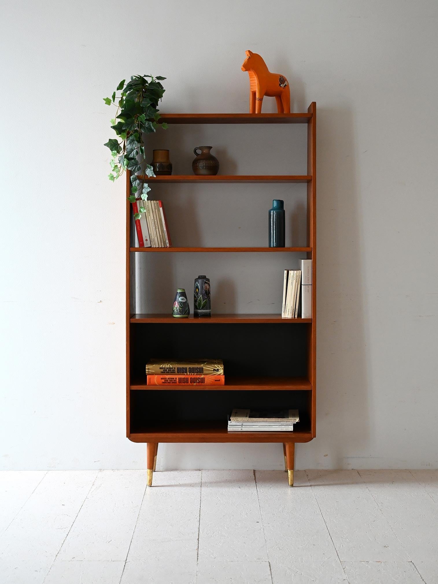 Vintage teak bookcase with metal feet and partially black bottom, featuring sloping profile and raised base.

Scandinavian 1950s/60s design with teak frame and tapered feet with gold metal finials. The sloping side profile lends dynamism to the