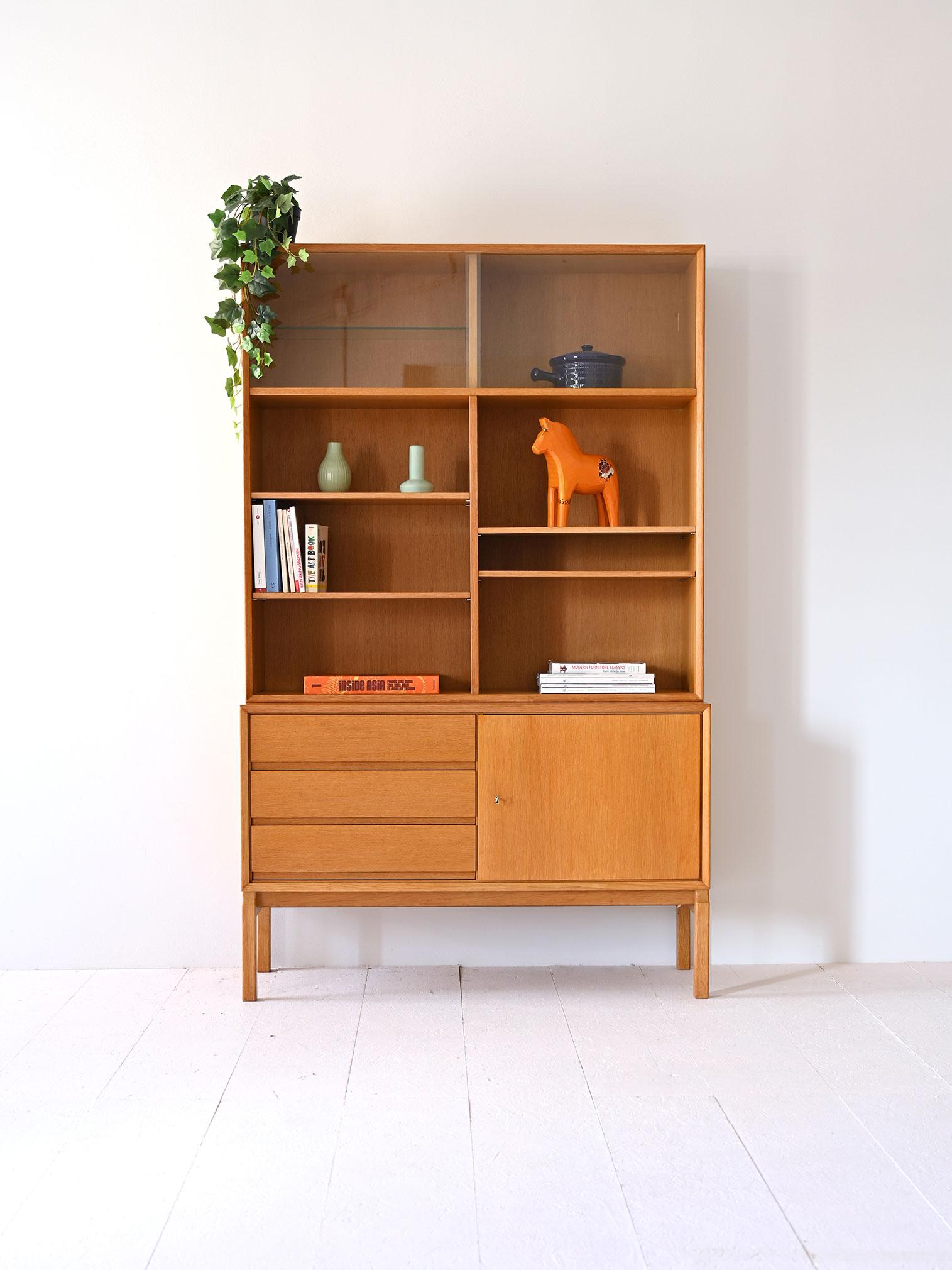 Vintage Bücherregal bestehend aus zwei Modulen, der untere Teil ist ein kleines Sideboard mit einer Tür und Schubladen, während der obere Teil eine Struktur mit offenen Regalen und einer Vitrine ist.

Die klaren Linien und der helle Farbton der