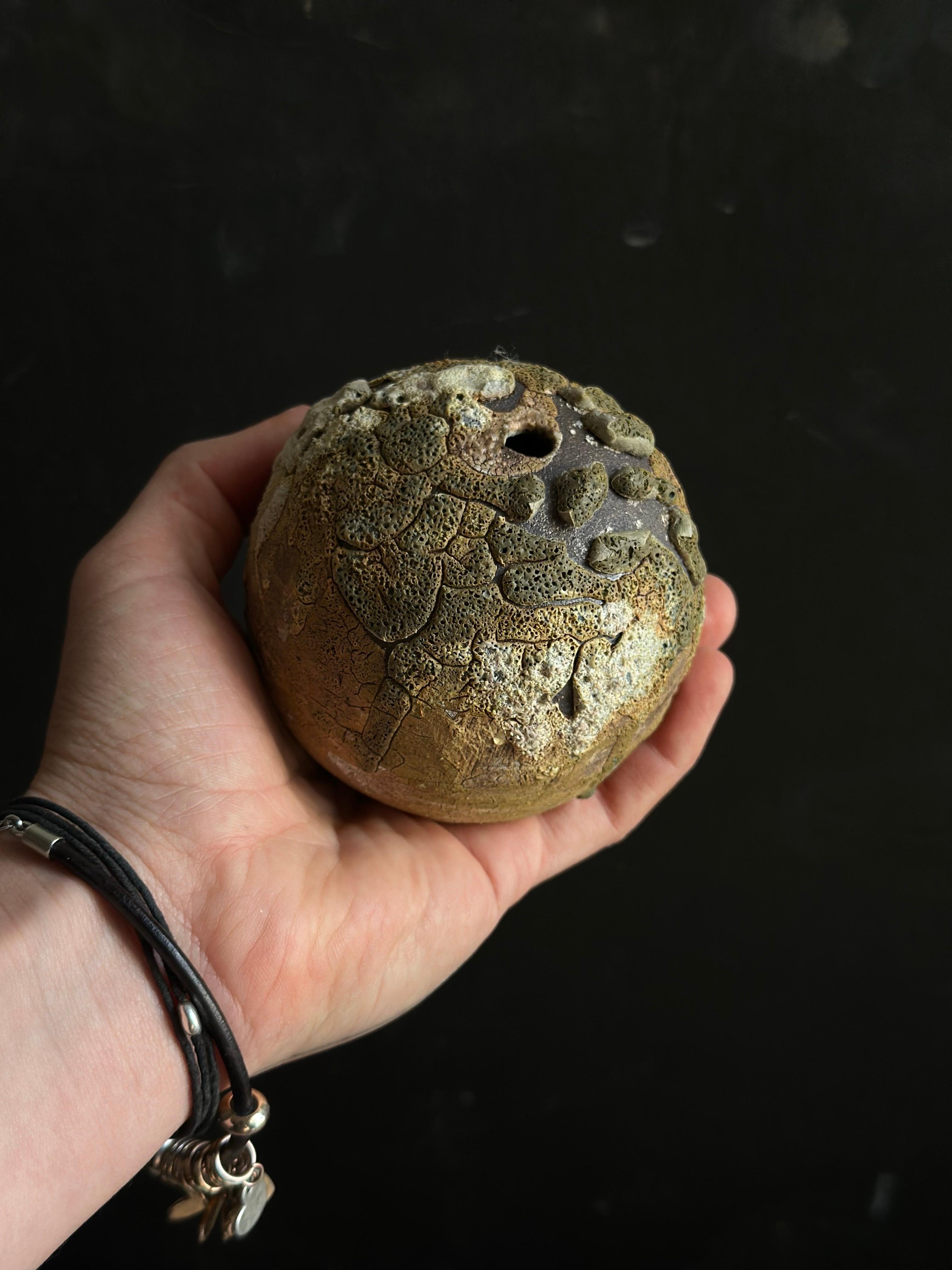 A unique ceramic bud vase featuring a richly textured surface reminiscent of lichen-covered stones. The earthy tones, combined with pops of green and ash-like accents, give this piece a natural, weathered appearance. Perfect for minimalist or