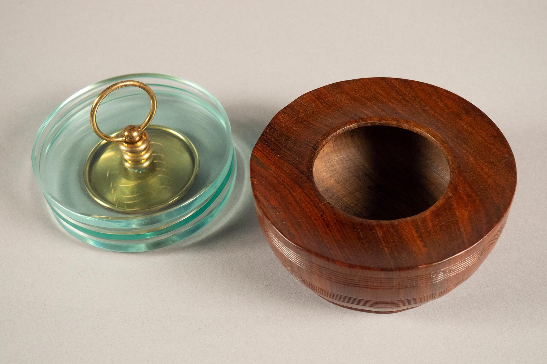 Round wooden box, topped by a glass lid comprising three stacked glass discs, held together by a brass handle.