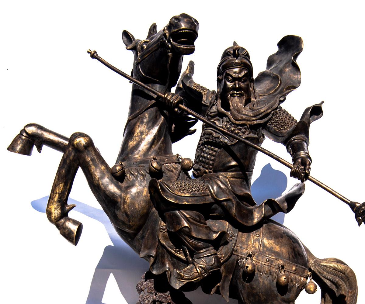 This life-size bronze with gold highlights of the Chinese General Guandi upon horseback brandishing his sword as his beard flies in the wind. The horse is resting upon the ground with bonzi like tree below the front hooves as he rears back in an