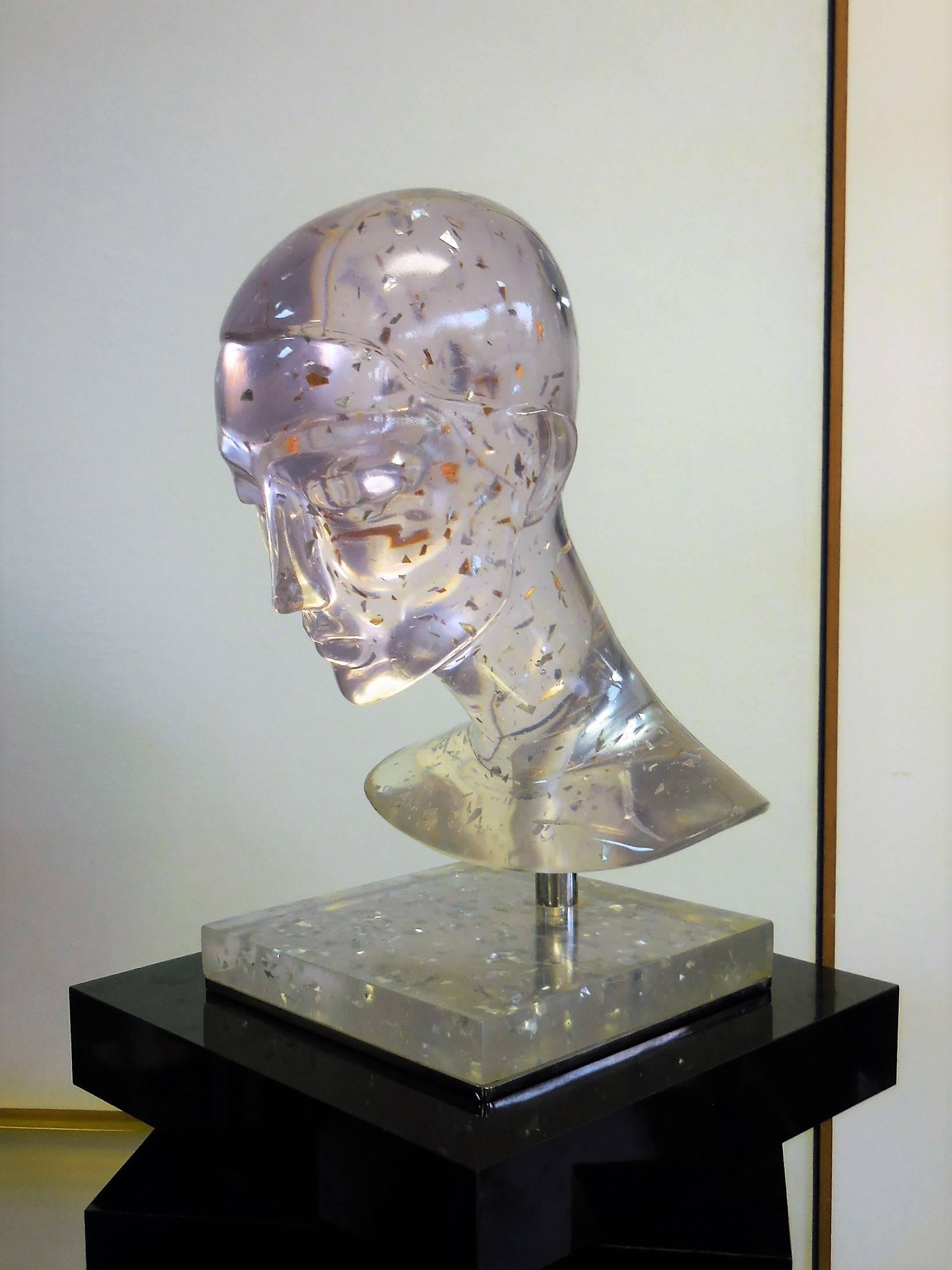 A large sculpture in clear resin with metal inclusions. Depicts the stylized head of a woman with Classic features. The head rest on a chrome base, included is a revolving black laminate pedestal. Sculpture without the pedestal measures 15.5
