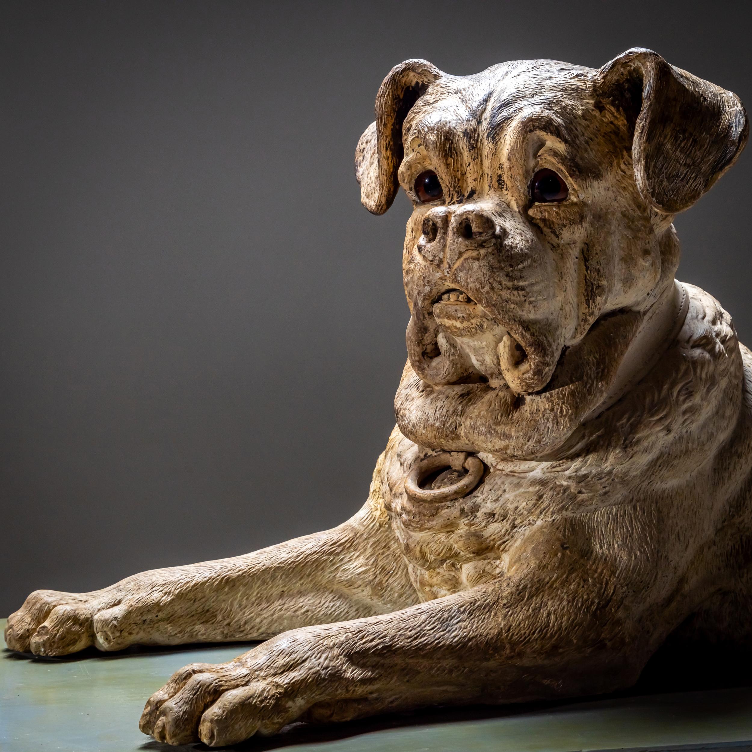 A naturalistically crafted, life-size dog sculpture made of painted ceramic. The dog is depicted in a reclining yet alert posture. Its brown glass eyes enhance the lifelike appearance.

The figure represents a German Boxer with distinctive features