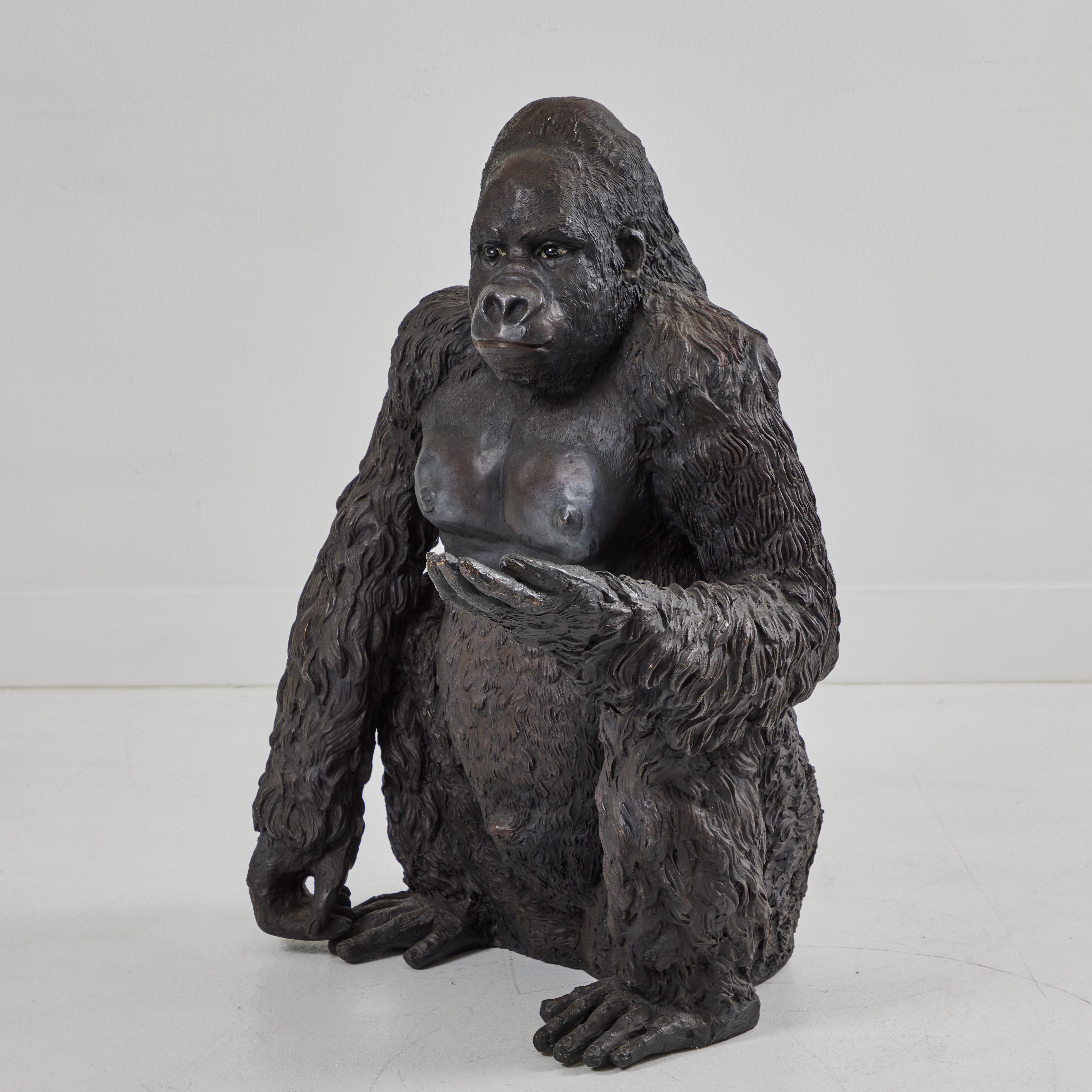 Dieser bronzene Gorilla sitzt aufmerksam mit ausgestreckter linker Hand und entspanntem rechten Arm auf dem Boden. Die schwarzen Glasaugen reflektieren das Umgebungslicht und verleihen diesem Kerl ein mitfühlendes und lebensechtes Gesicht. Die