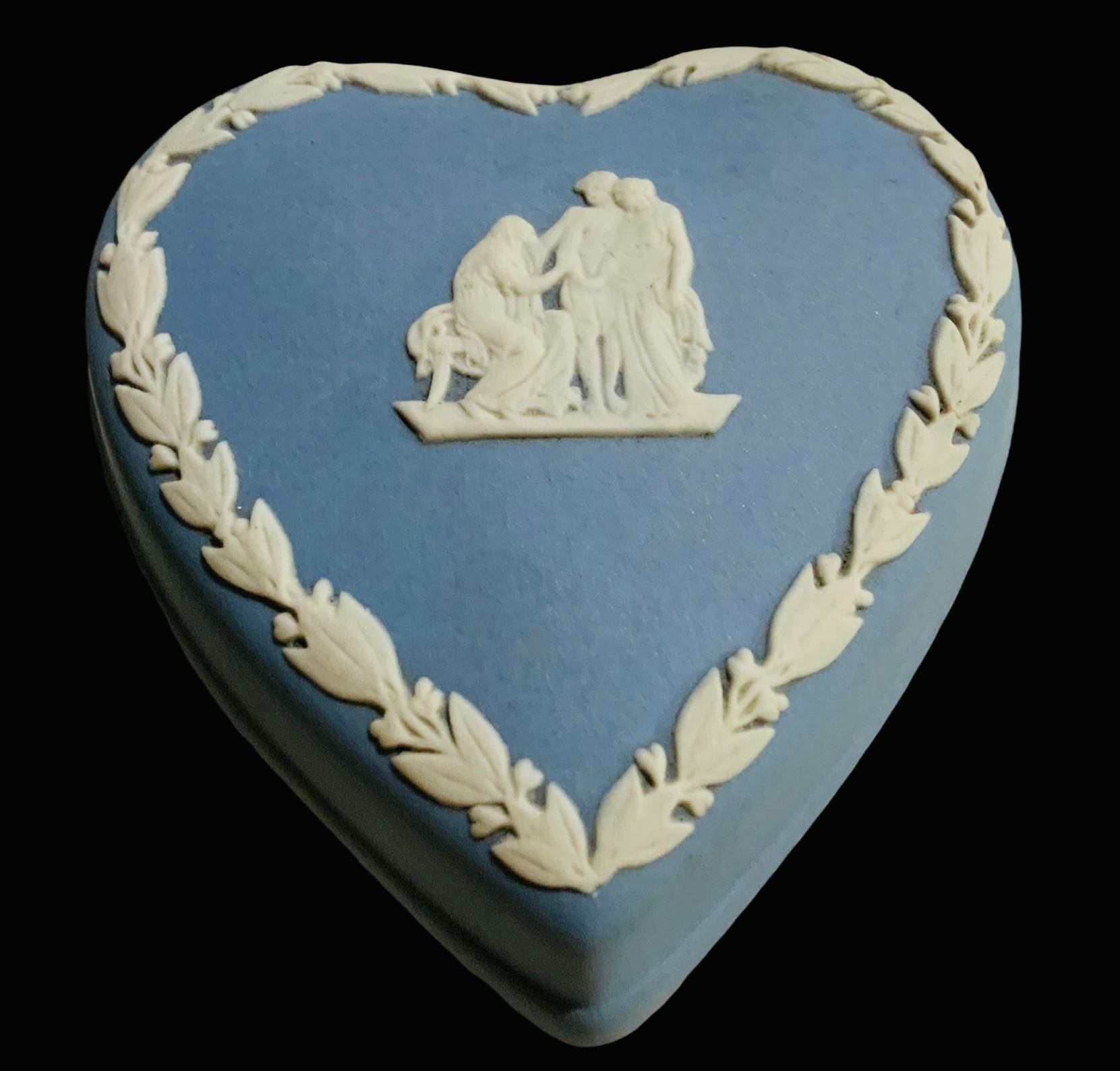 This is a light blue Wedgwood heart shaped small lidded box. The top of its lid is decorated with a Greek or Roman relief scene of a woman with a cape covering her head sat in small bench talking or giving a blessing to a young couple that are