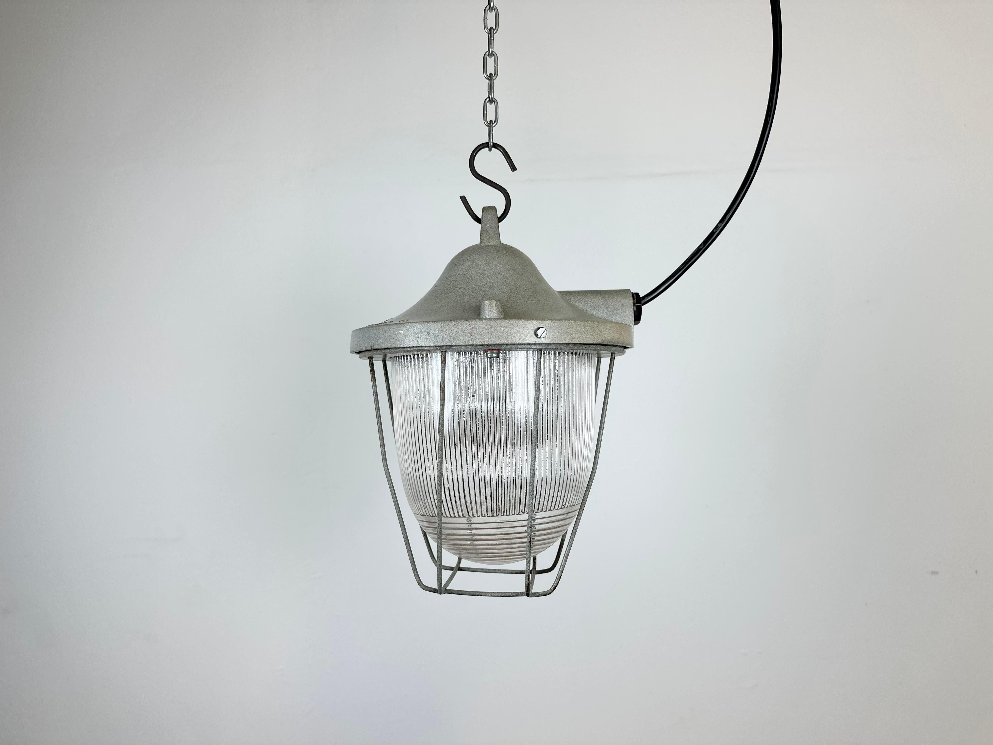 Light grey industrial lamp made by Polam Gdansk in Poland during the 1960s. It features a cast aluminium body, an iron cage and striped glass. The porcelain socket requires E 27/ E 26 lightbulbs. New wire. The weight of the lamp is 2,5 kg.
 
