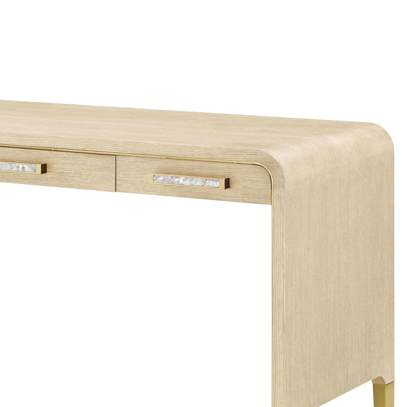 This desk blends timeless form with luxurious detail. Its elegant waterfall silhouette is softened by rounded corners and wrapped in a light wood finish. Polished brass trim outlines the frame, while mother-of-pearl inlaid pulls add a subtle,