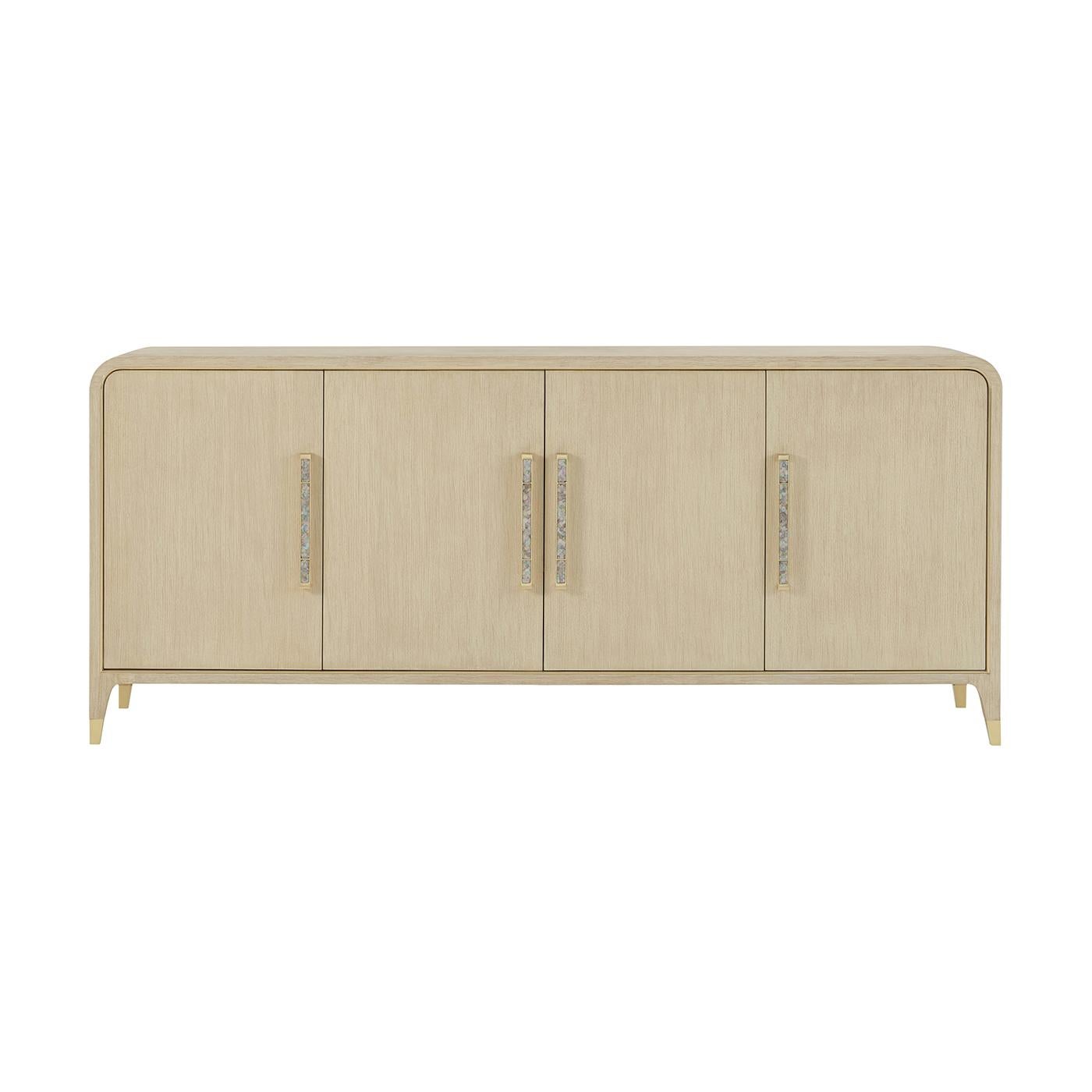 Shown in a light wood martini finish, its four sleek doors are framed with soft curves, outlined in gleaming brass, and accented with elongated pearlescent inlaid hardware.

Tapered brass-capped feet elevate its sculptural presence, while the