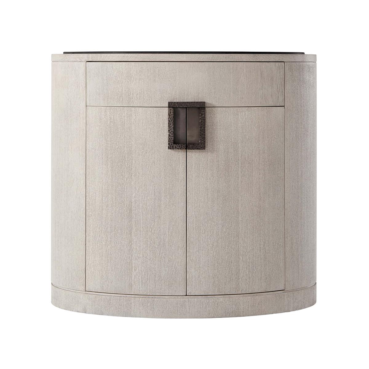Light oak oval nightstand in a brushed oak gowan finish, the oval basalt inset top, with a hammered metal handle with a matte tungsten finish, a frieze drawers above a two-door cabinet enclosing an adjustable shelf.

Dimensions: 30
