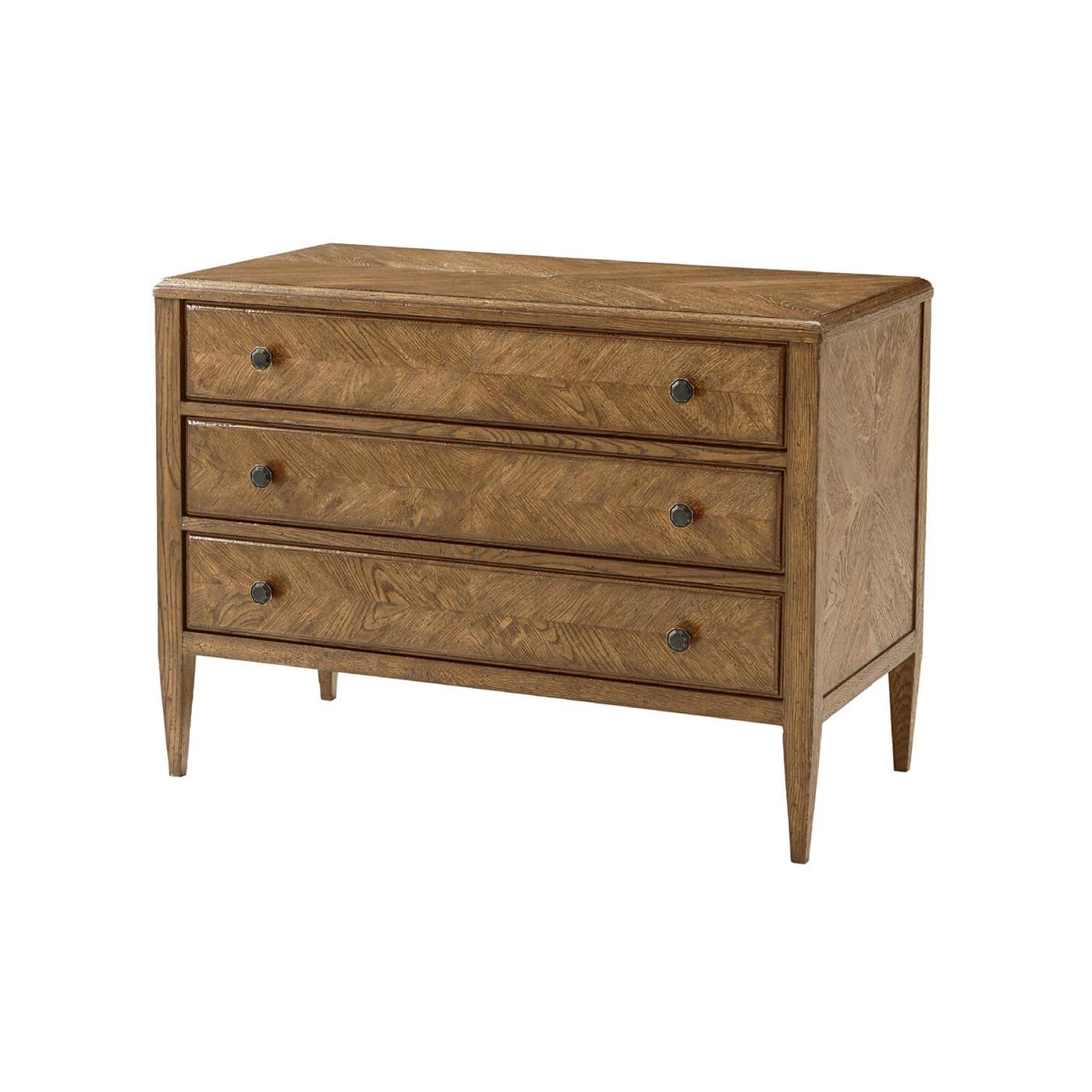 A light oak parquetry chest of drawers in dawn finish. It has mirrored herringbone oak parquetry drawers, classically tapered legs, and six Verde Bronze finish handles. 
Shown in dawn finish
Dimensions: 38
