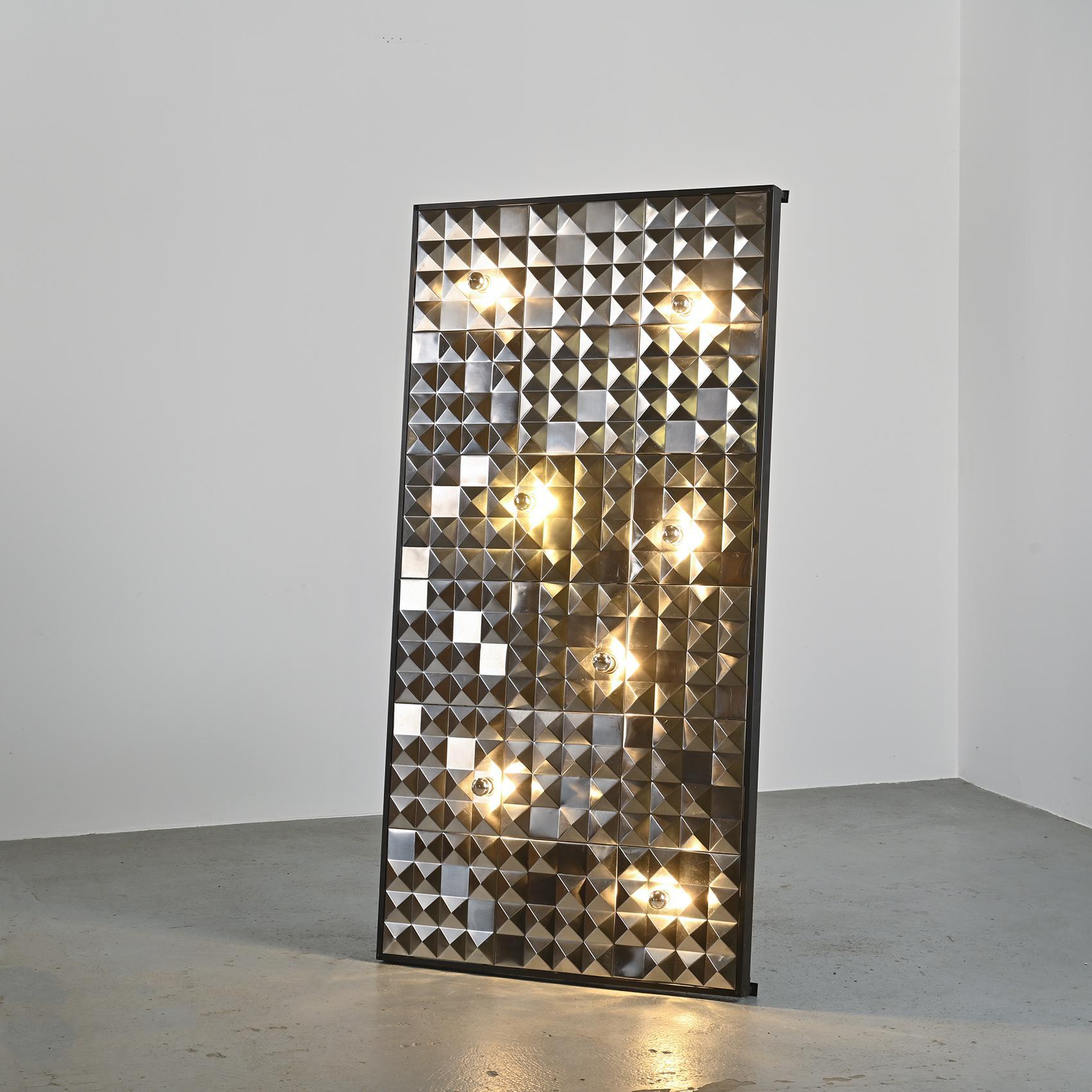 Satz von achtzehn dekorativen Paneelen aus poliertem Edelstahl mit Rautenmuster. Auf einem Holzrahmen montiert und von sieben Glühbirnen mit versilberter Kappe beleuchtet, wird das Werk zu einer echten Lichtskulptur.

Durch Reflexion und Brechung