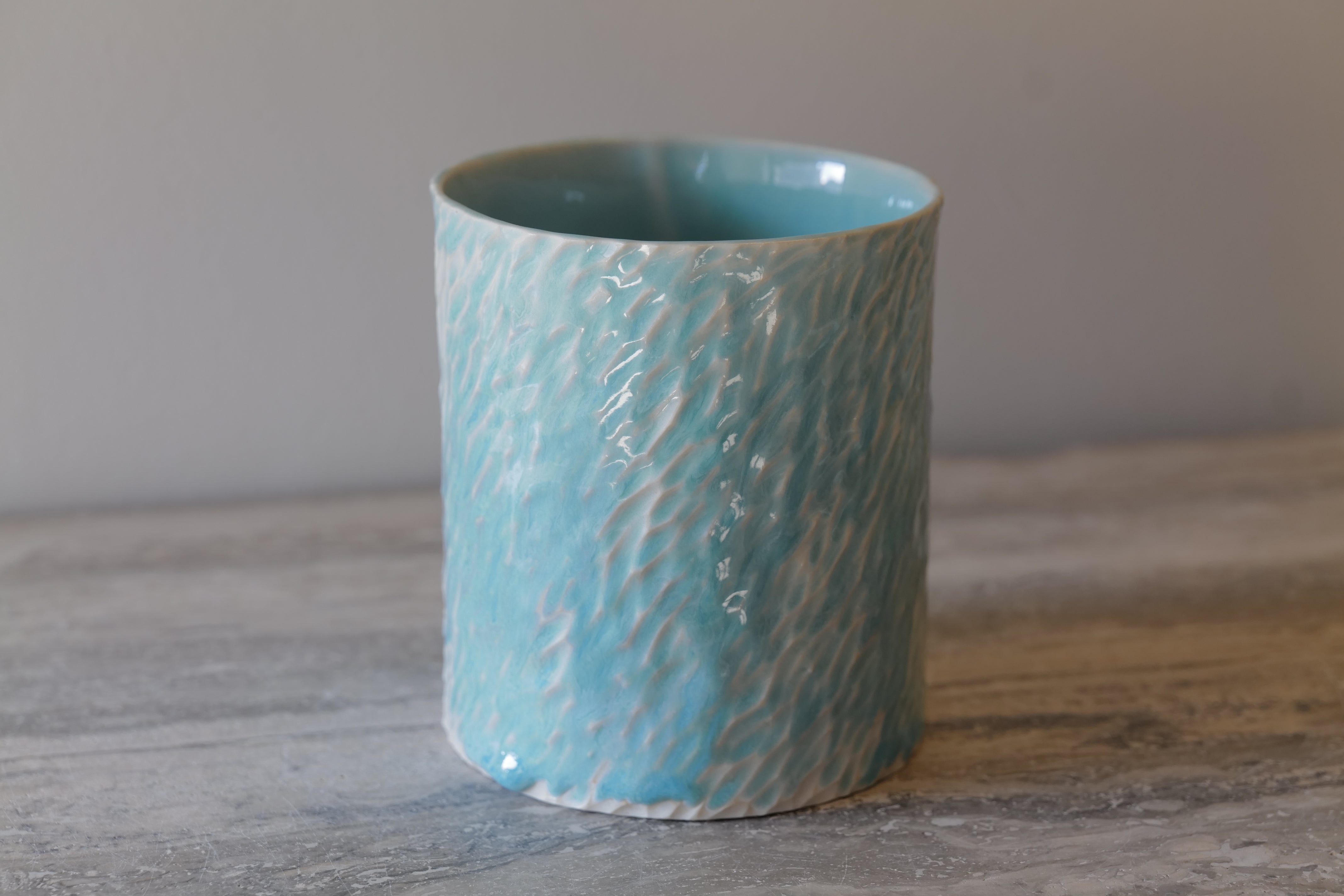 Das Light Turquoise Carved Cylinder Vessel ist ein heiteres und modernes Stück, das die zarte Schönheit von Wasser und Licht einfängt. Aus feinem Porzellan gegossen und sorgfältig von Hand geschnitzt, weist seine Oberfläche sanfte, wellenartige