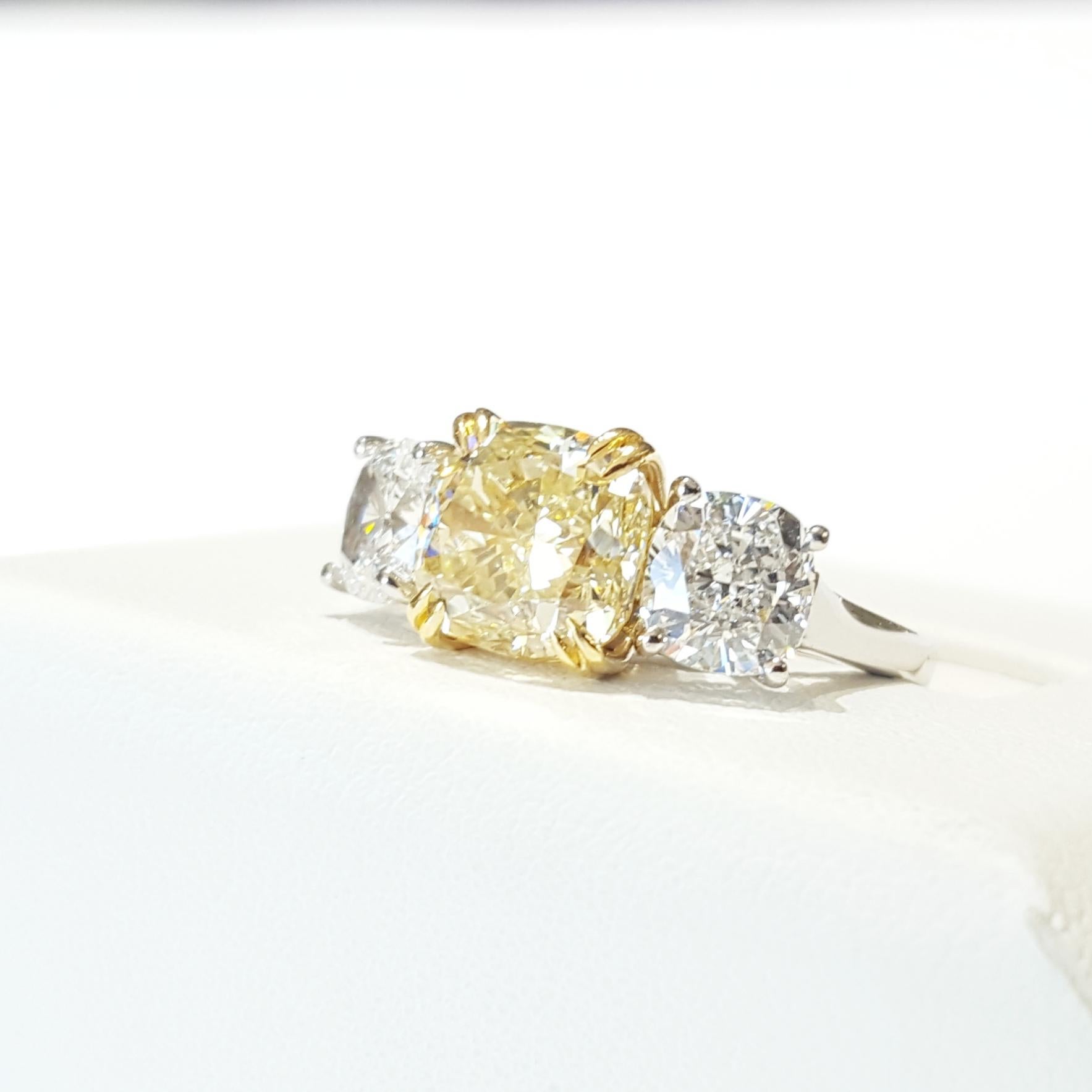 A platinum ring set with 1 light Yellow Cushion Cut diamond and 2 White Cushion Cut diamonds. The center diamond weighs 3.02ct, measures 7.91 x 7.71 x 5.72mm is light yellow and VS2 clarity. The side diamonds, are G color Si1 clarity and weigh