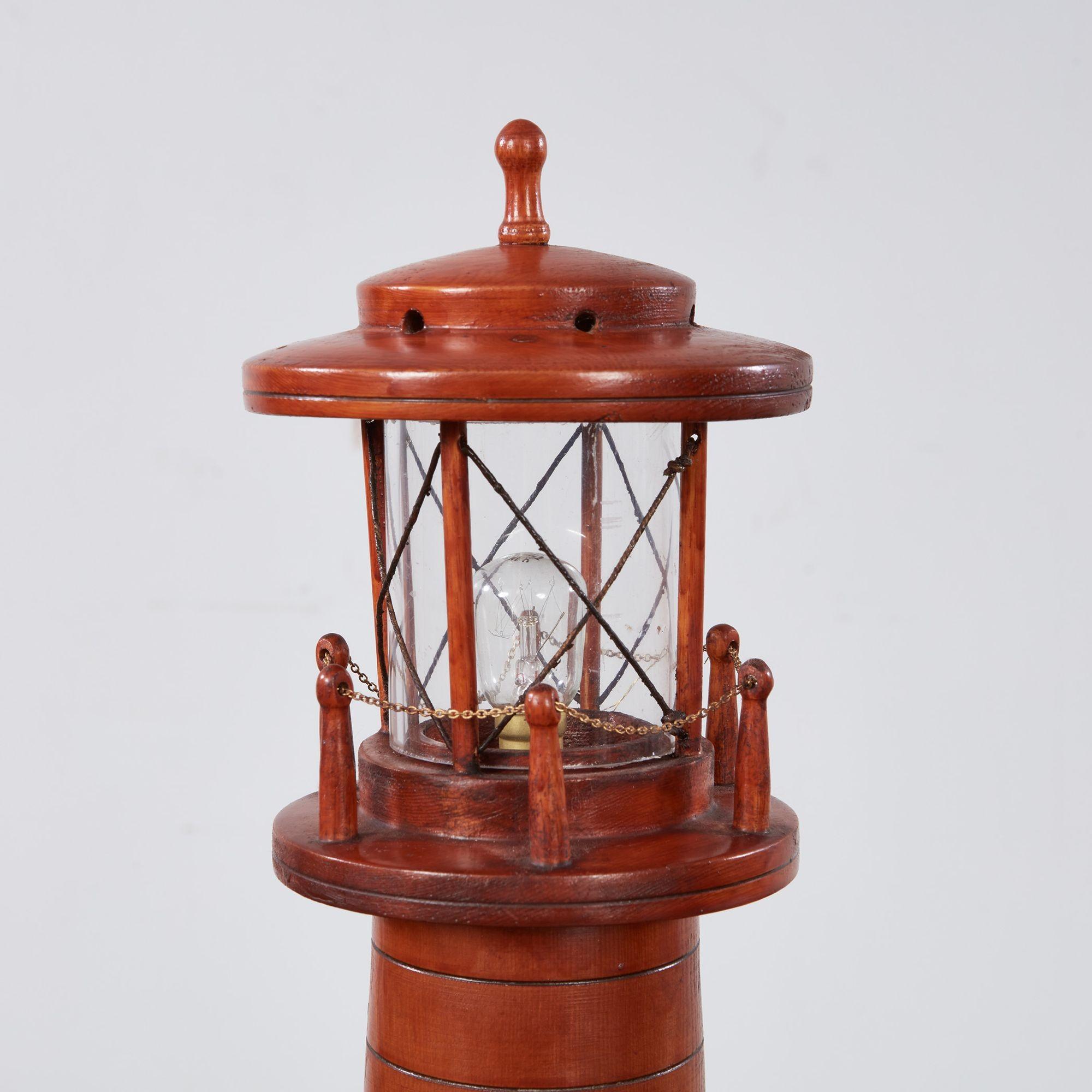 Lámpara de sobremesa faro de madera tallada con paredes cónicas y anilladas sobre una base torneada, rematada por una carcasa de luz de cristal rodeada por una barandilla de postes y cadenas y coronada por un tejadillo y un remate.  Contiene una luz
