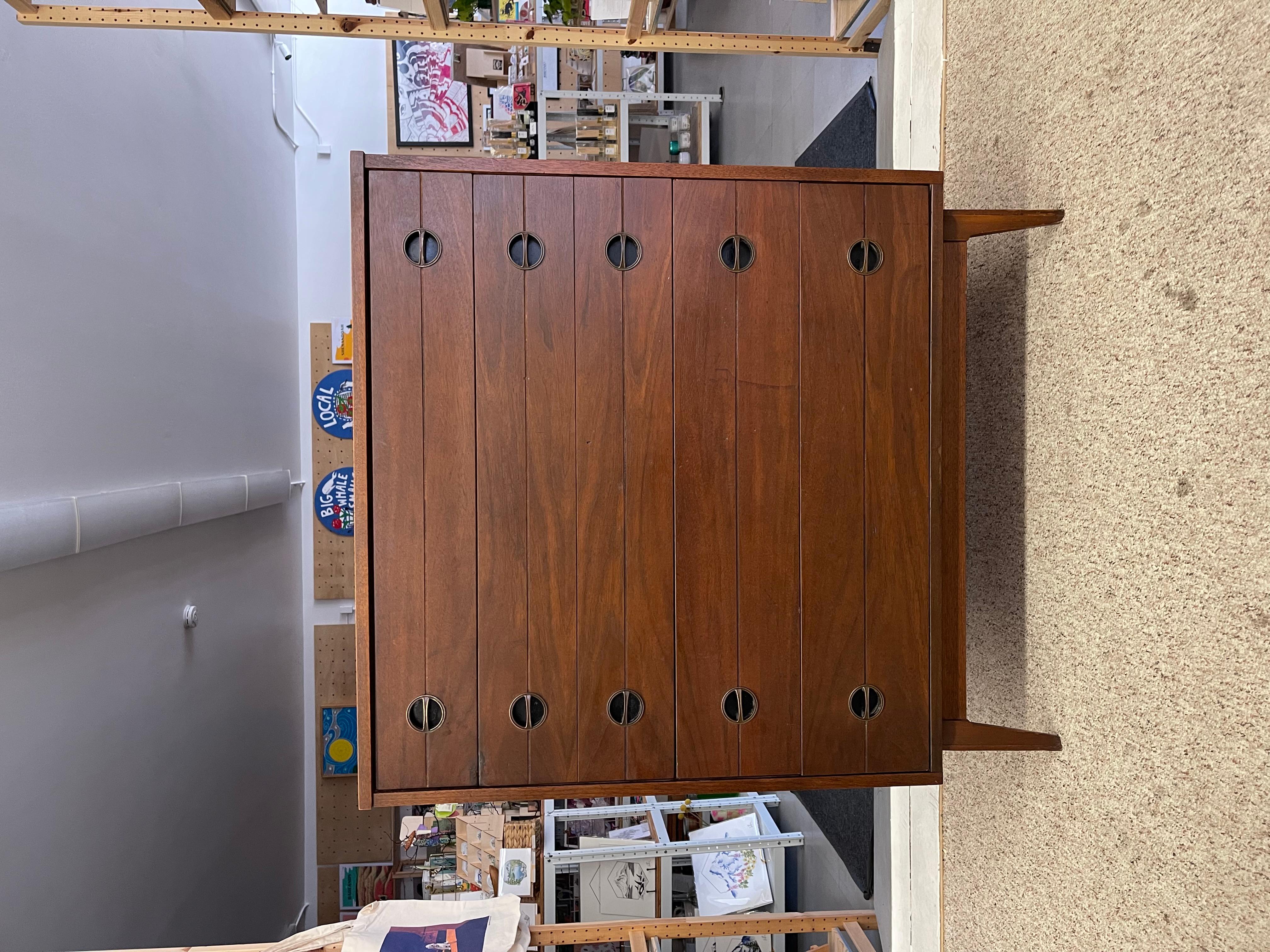 La commode haute vintage Mid Century Modern, datant très probablement des années 1960, est fabriquée à partir de noyer massif possible avec une finition chaude de couleur brun moyen qui met en évidence les motifs de grain naturel dans l'ensemble.
La