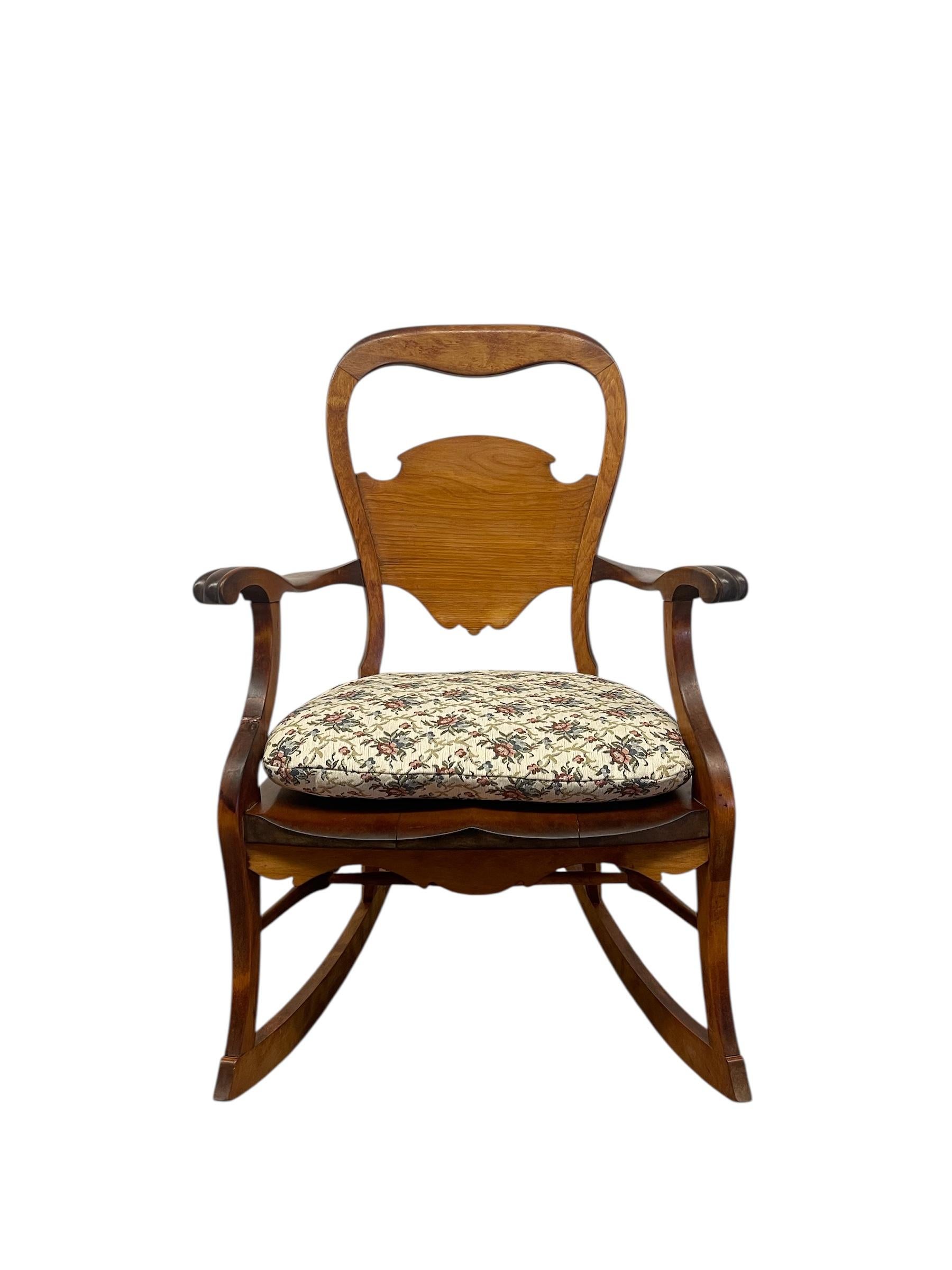 Questa probabile sedia a dondolo antica in stile vittoriano presenta una struttura in legno massiccio scolpito con curve fluide e un pannello sagomato sullo schienale. I braccioli sono delicatamente intagliati con dettagli a voluta alle estremità,